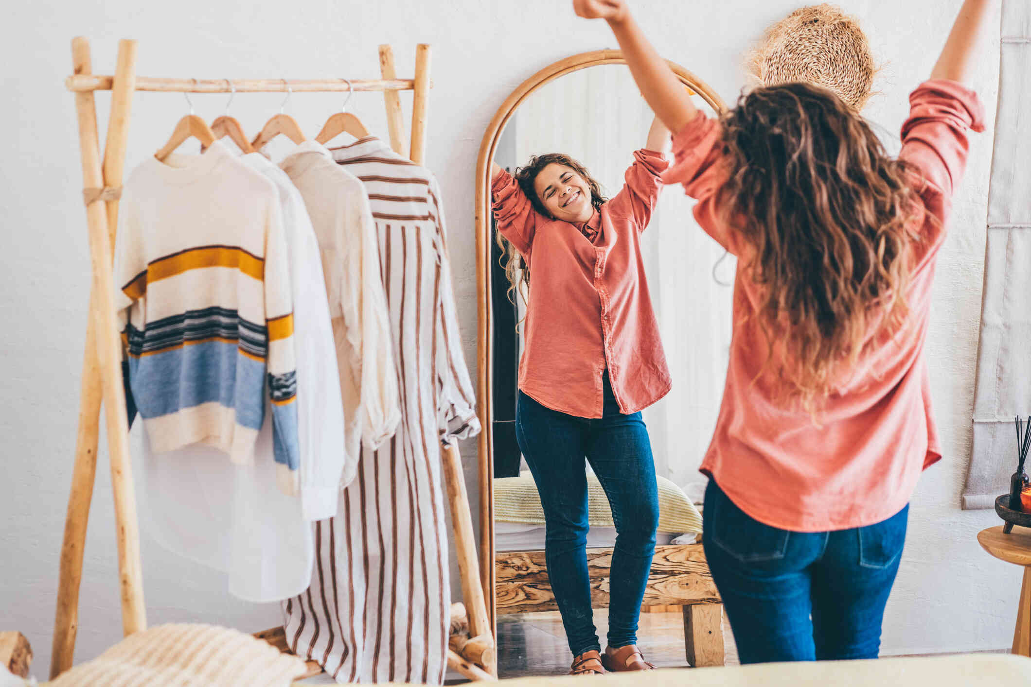 A woman in a button down shirt raises her arms happy as she looks in the full length mirror while standing next to a rack of clothes.