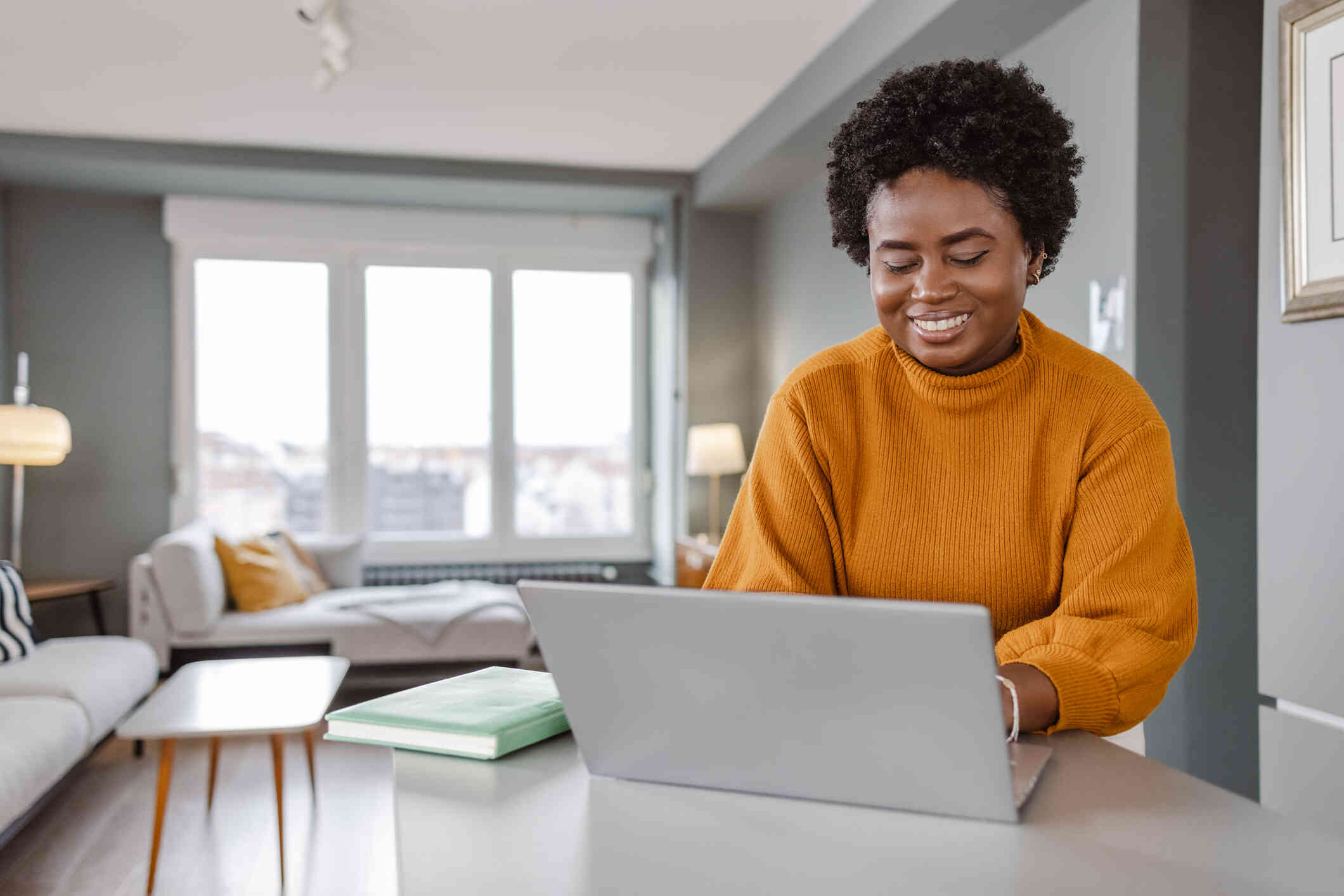 A woman in an orange sweater smiles at the laptop open on the table infront of her with her planner sitting next to it.
