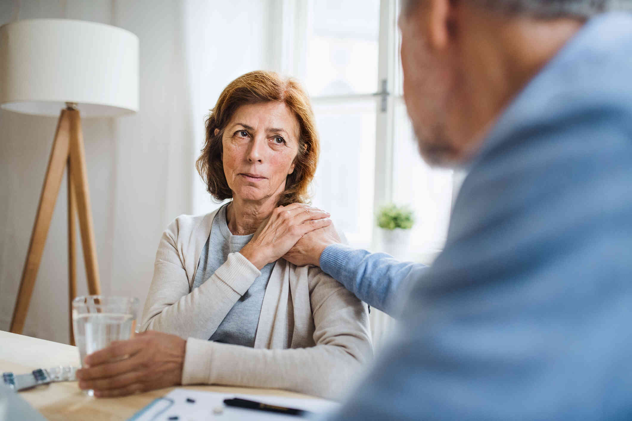 An older woman is looking distressed, sitting at a table while holding a glass of water, with a hand resting on her shoulder in a comforting gesture by a person seated across her, offering support.