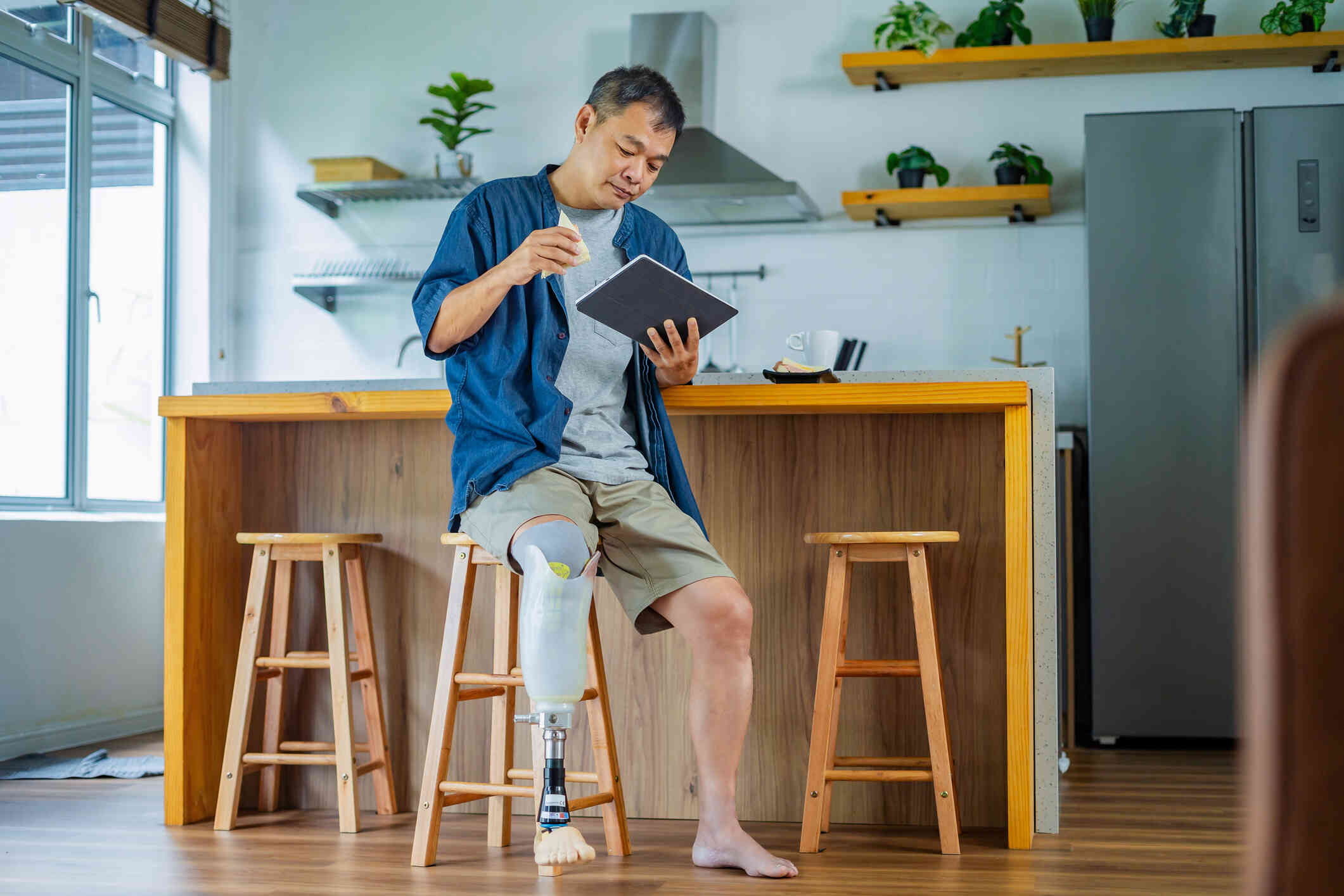 A man with a prosthetic leg sits on a stool at the breakfast bar in his home and holds a sandwhich in one hand while looking at the tablet in the other.