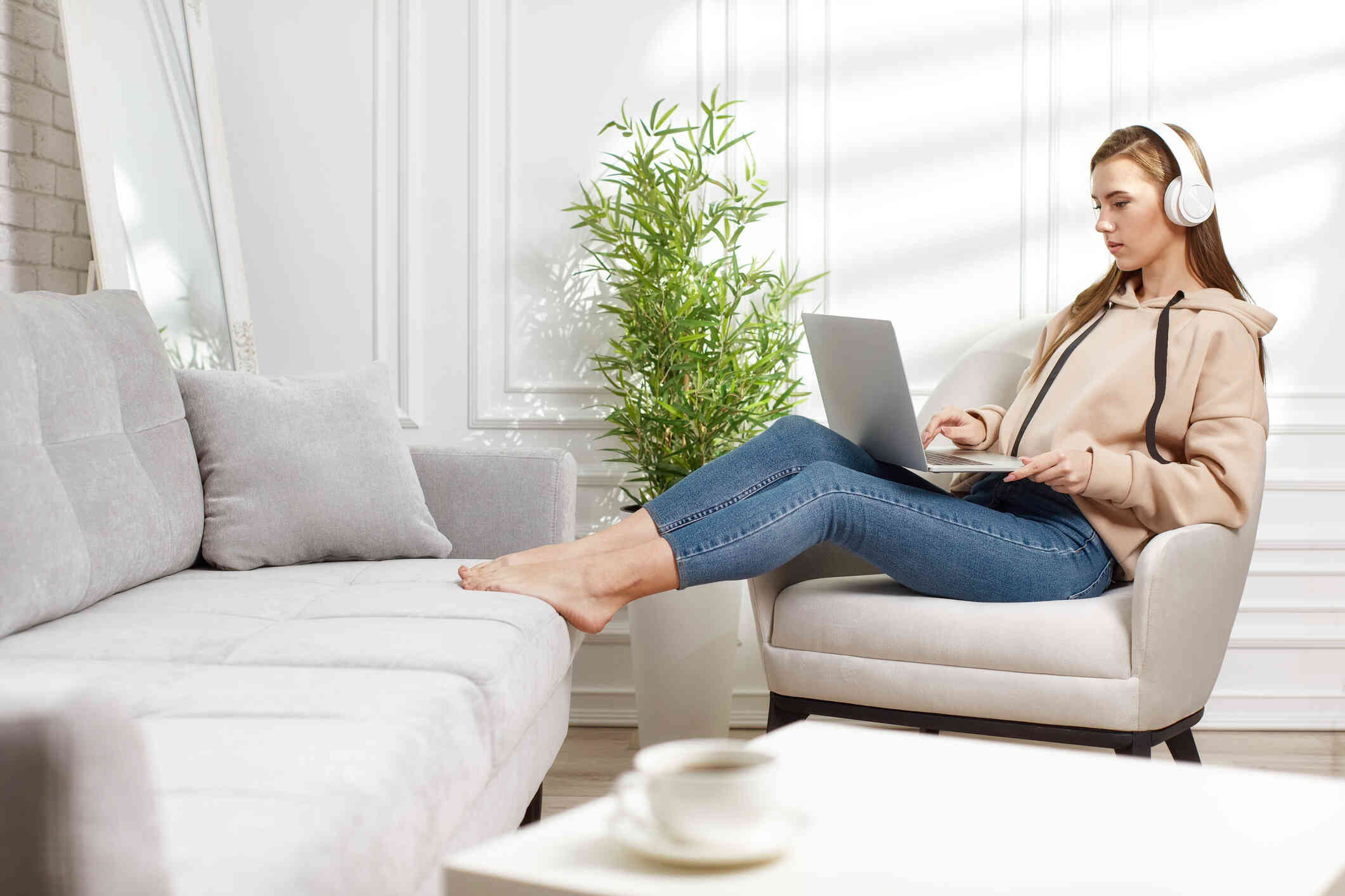 A woman sits in a chair in her home with her feet restin on the couch as she wears white headphonesand pooks at the laptop open on her lap.