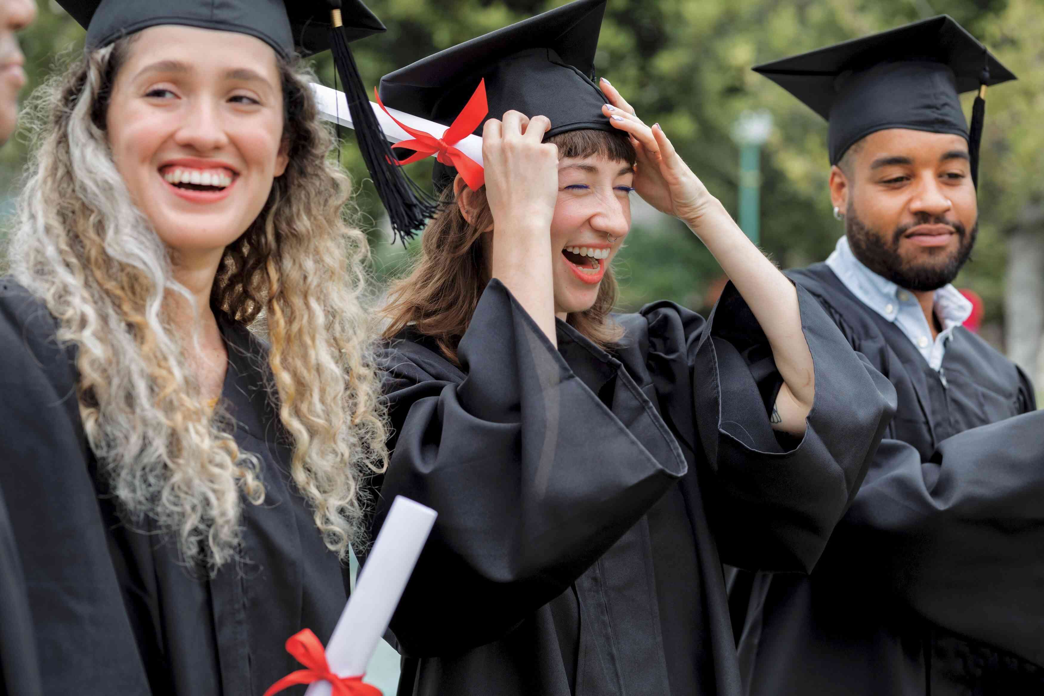 A young woman laughs and holds a diploma in her hand while grabbing her graduation cap