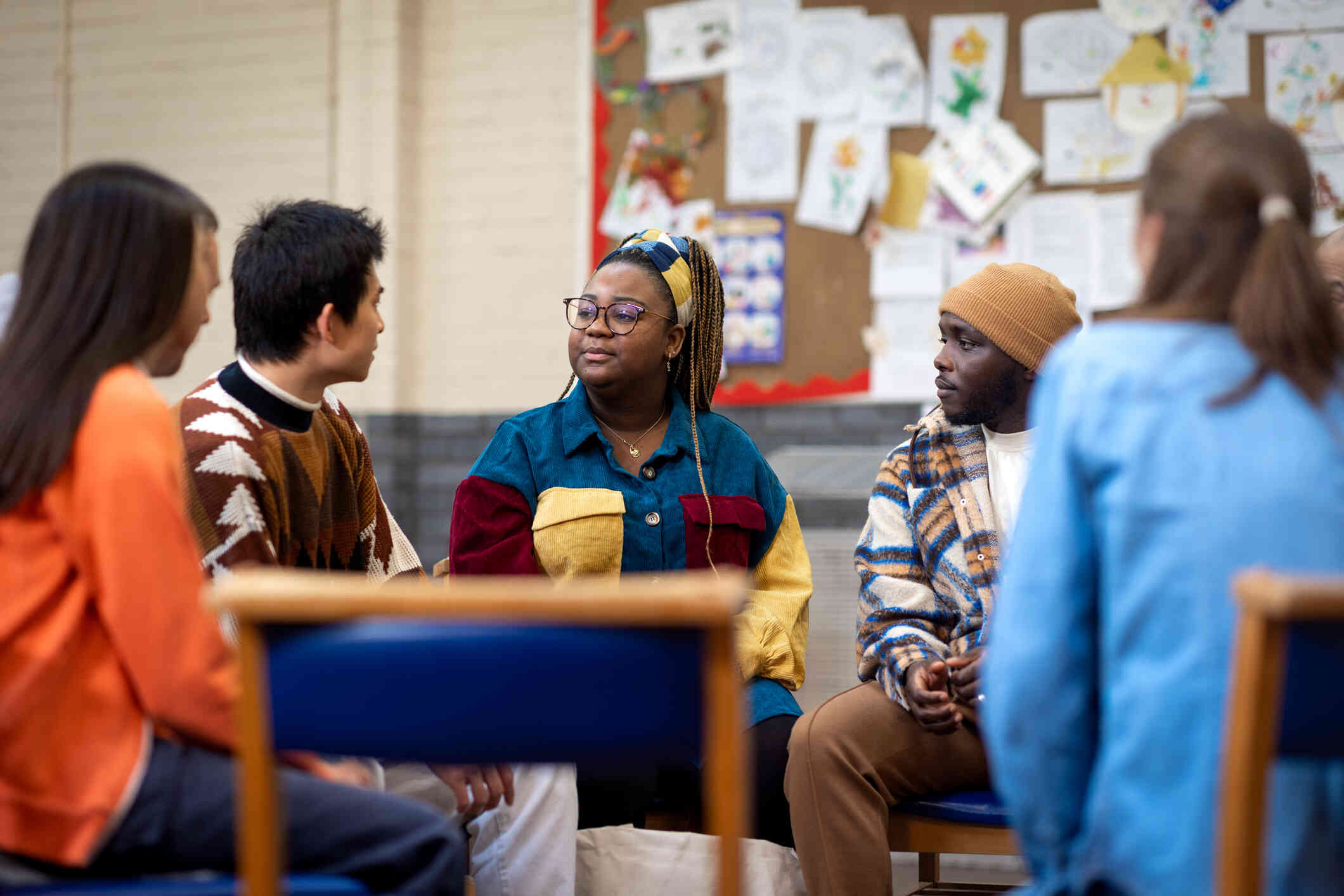 A group of young people sit together in a circle in a school building and a young woman with glasses, a headband, and braids looks at a young man who is speaking to the group.