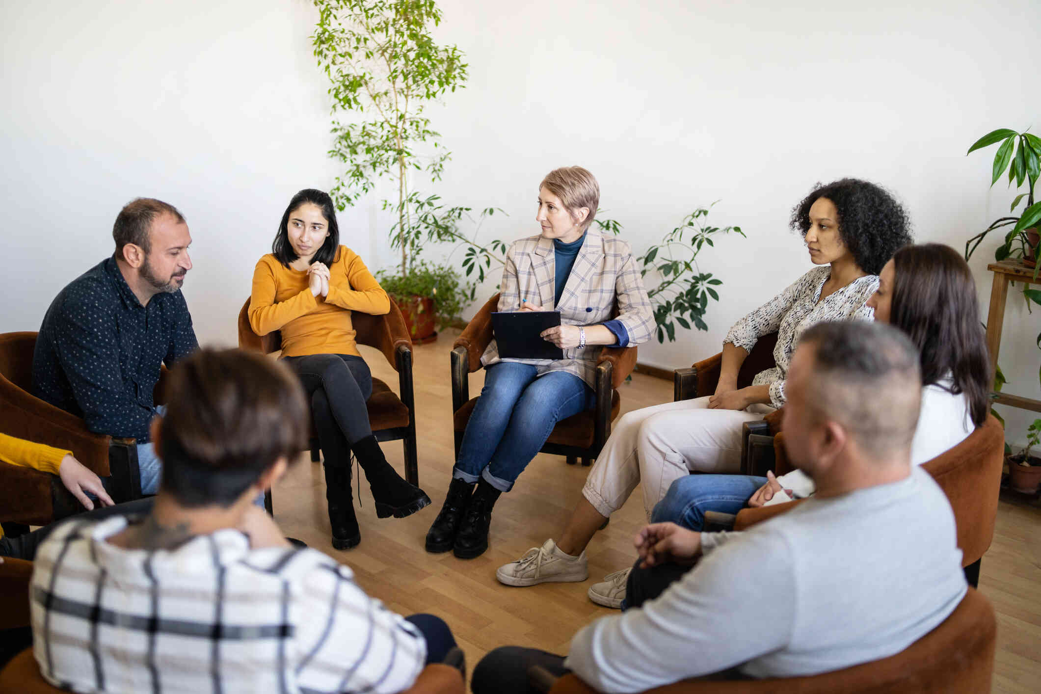 A group of adults sit in a circle in chairs and take turns talking during a support group meeting.