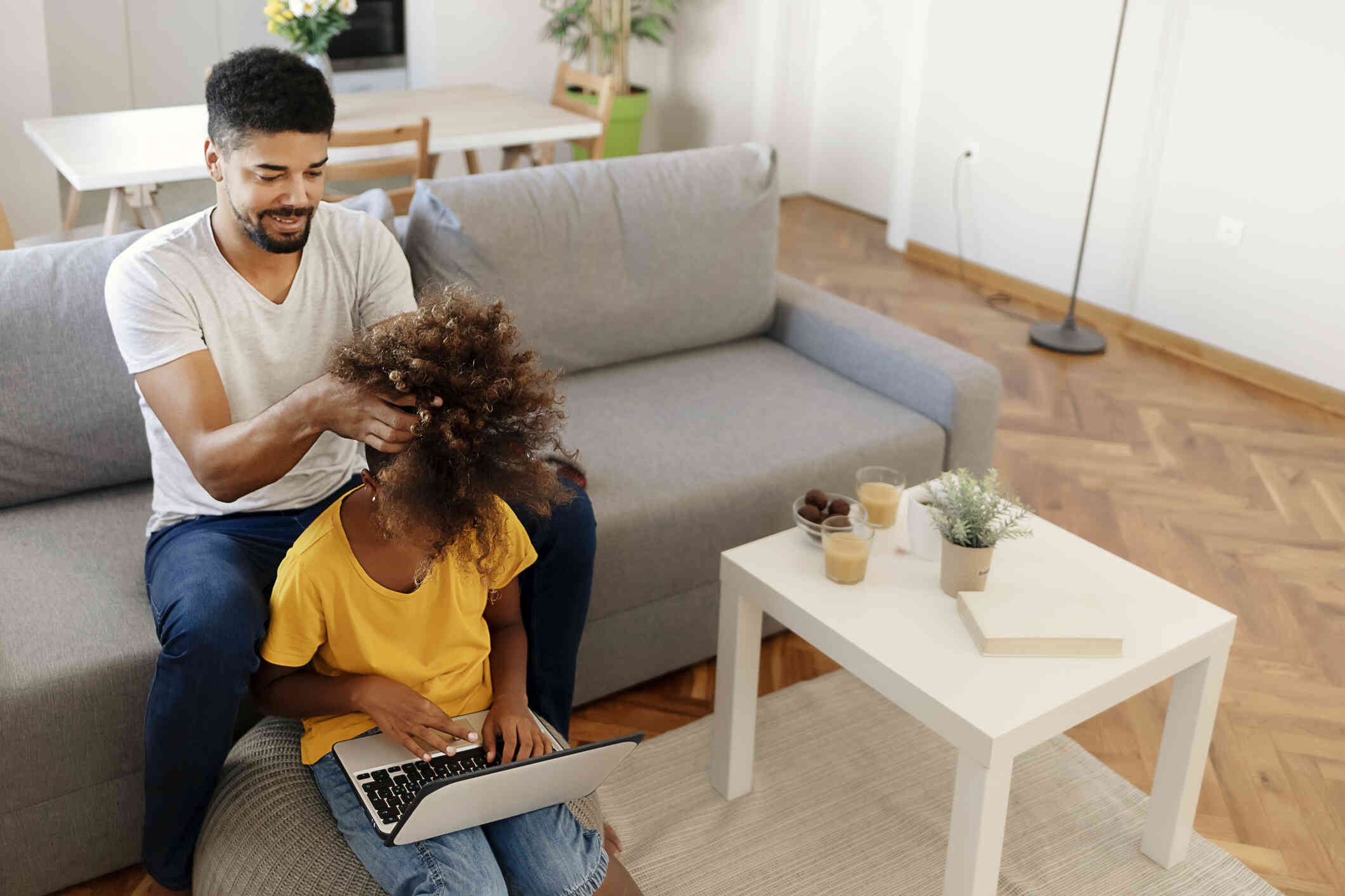 A girl in a yellow shirt sits on the floor with her laptop in her lap as her dad sits on the couch behind her and fixes her hair.