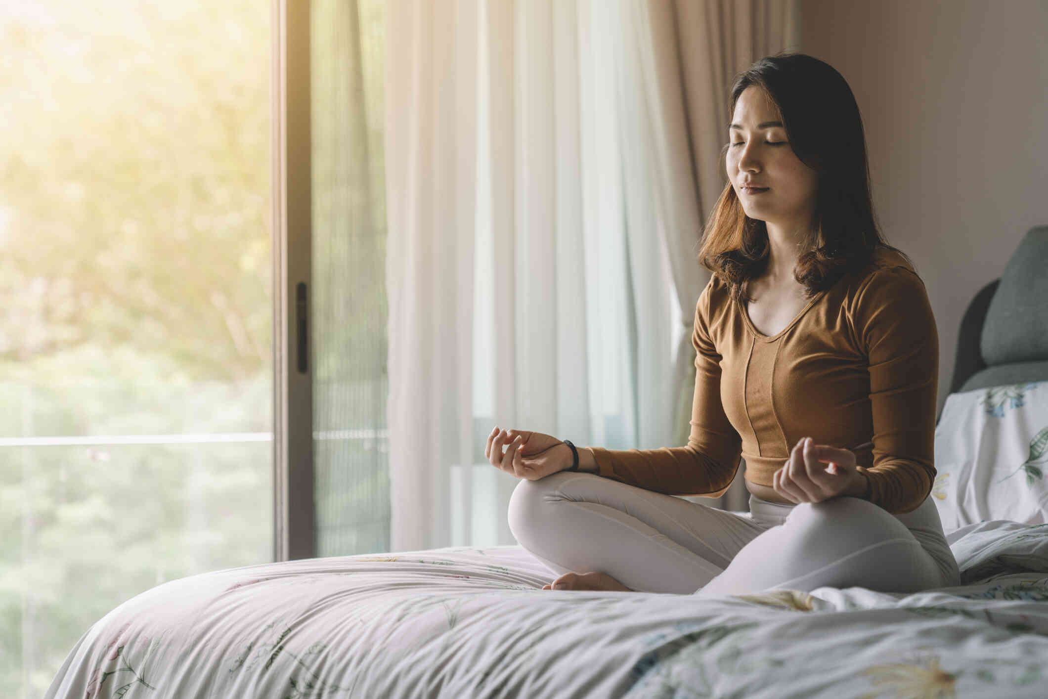 A woman is seated on her bed in a meditative pose.