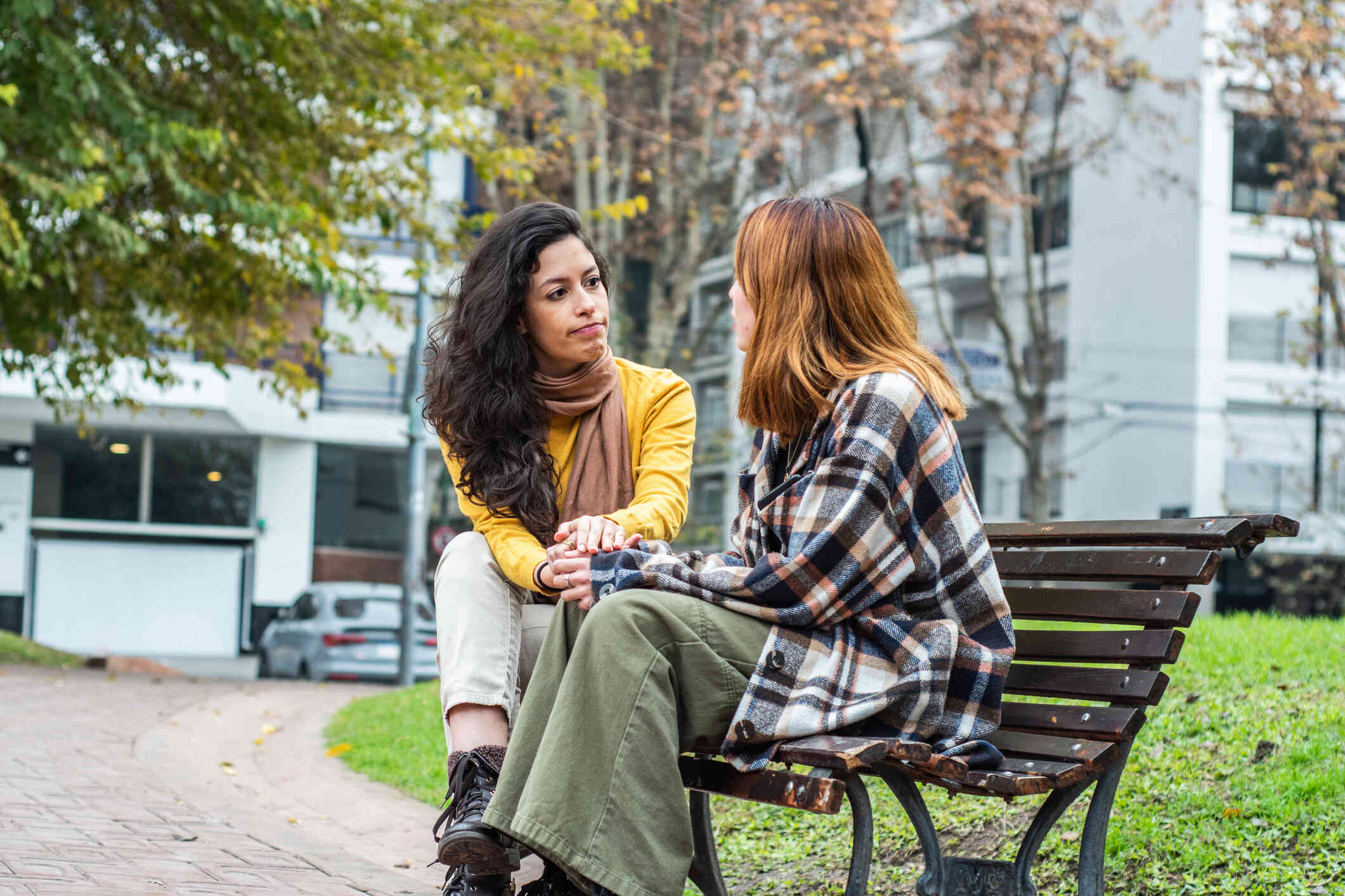 A female couple sit next to each other on a park bench while having a serious conversation.