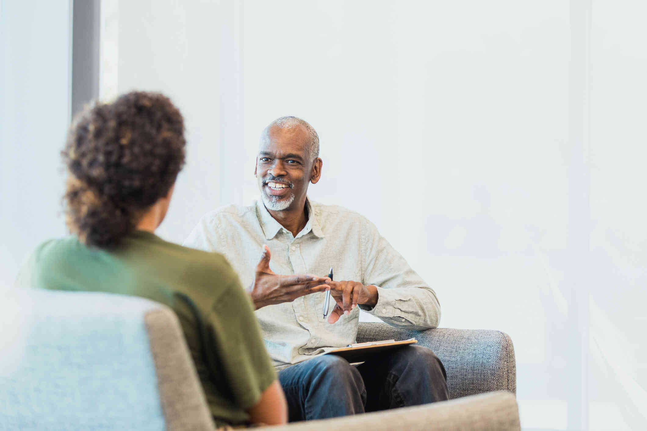 A male therapist sits across from his female patient and talks with a smile during a therapy session.