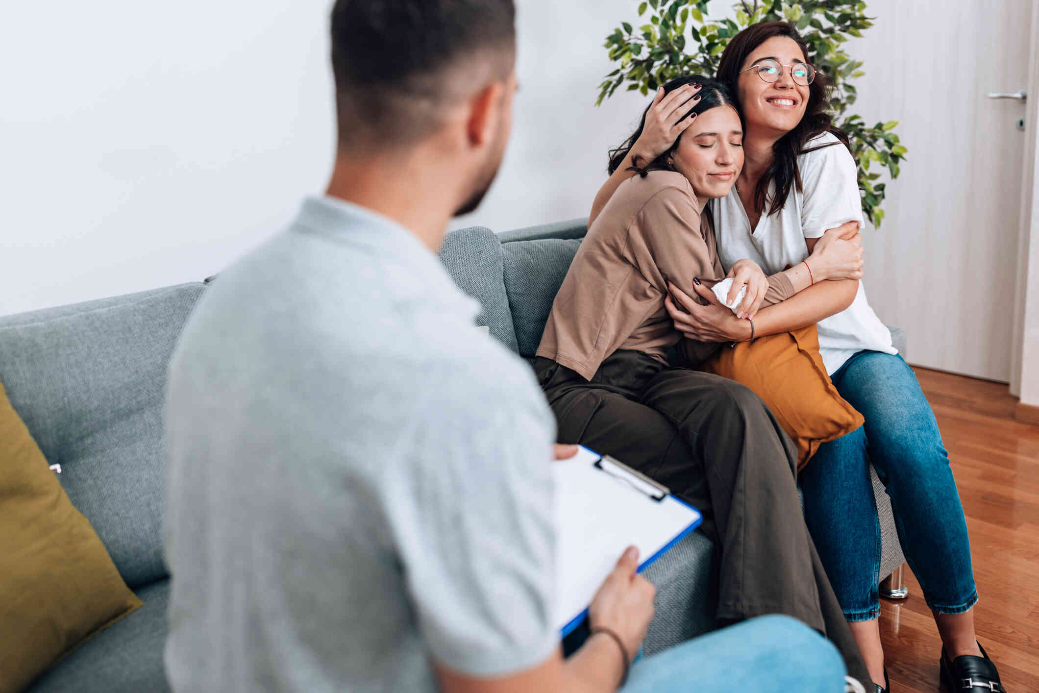 A young woman and her mother sit on a couch hugging with talking with a therapist wearing a blue shirt and holding a clipboard