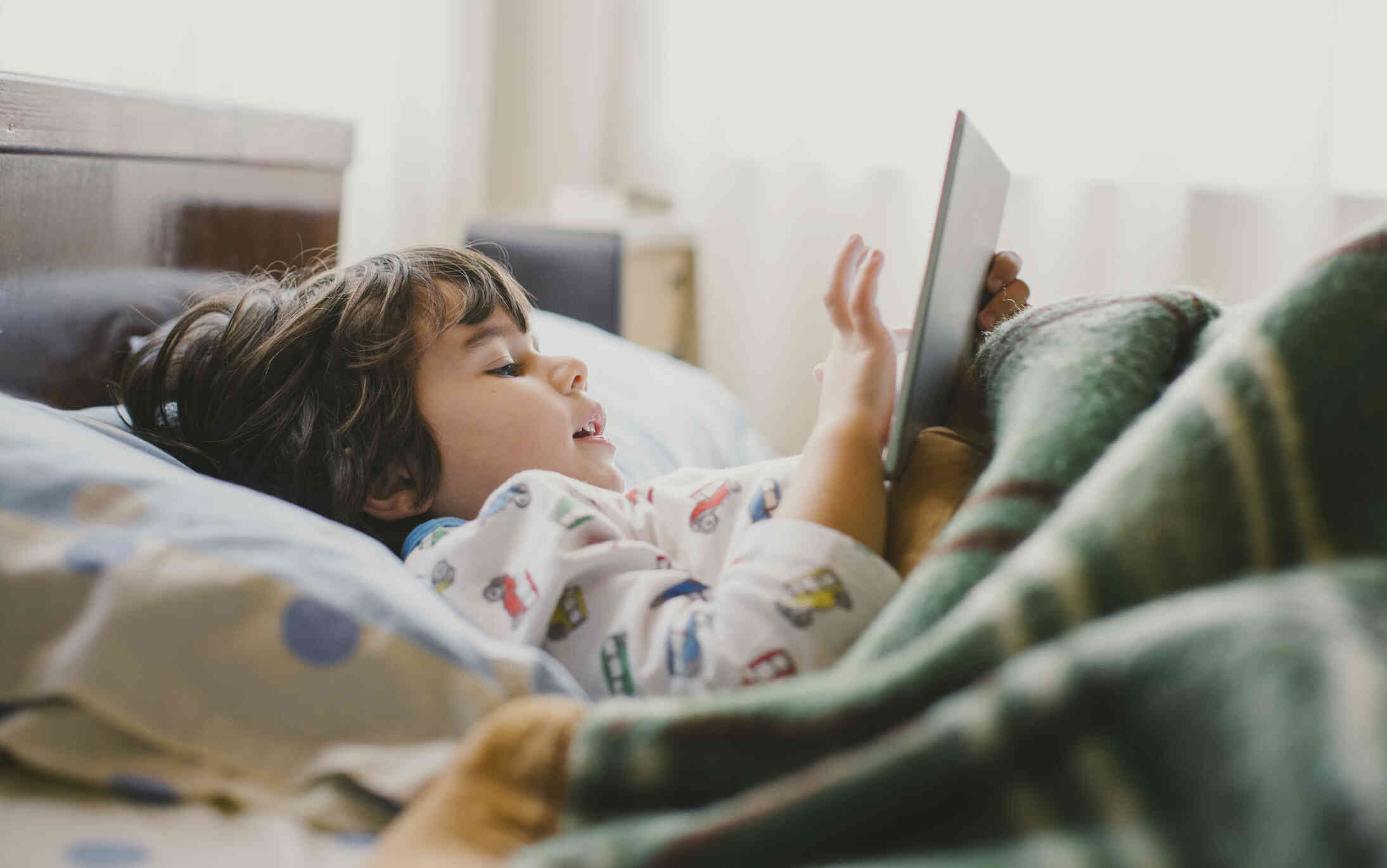 A little boy in pajamas lays on his back in bed under a blanket and uses his tablet while smiling softly.