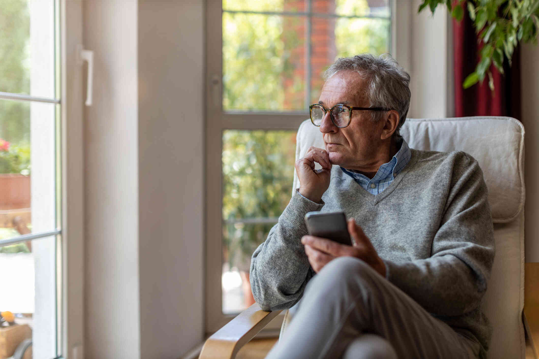 A middle aged man in a grey sweater sits in a chair in his home near a window with his phone in his hand as he gazes off deep in thought.