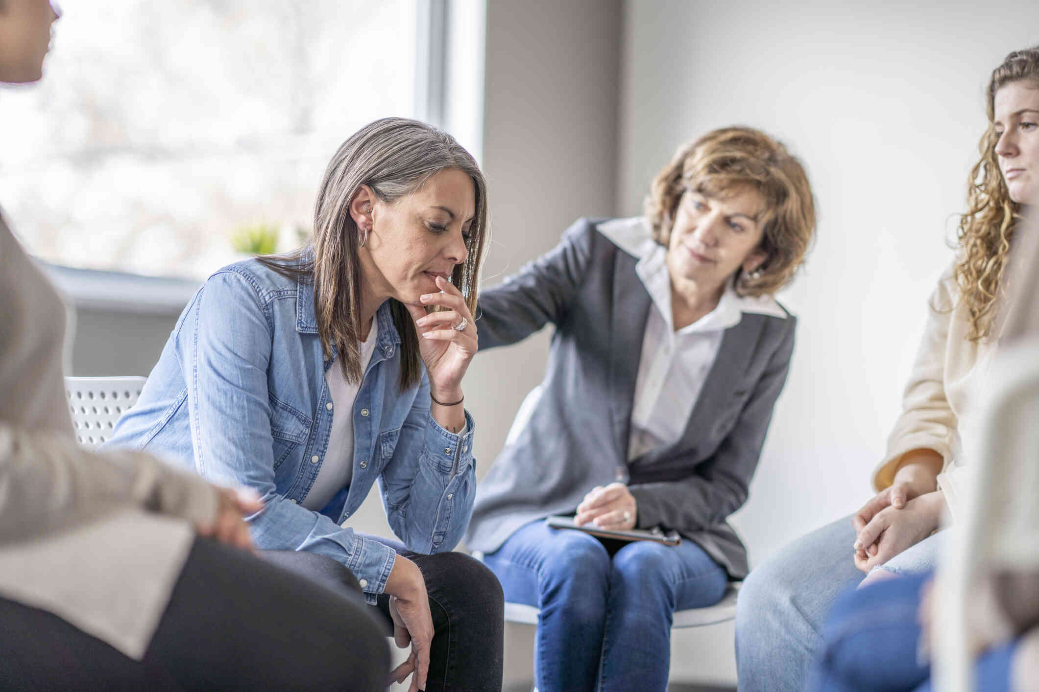 A group of adults sit in chairs in a circle as a female therapist reaches out to touch the shoulder of an upset woman during a group therapy session.