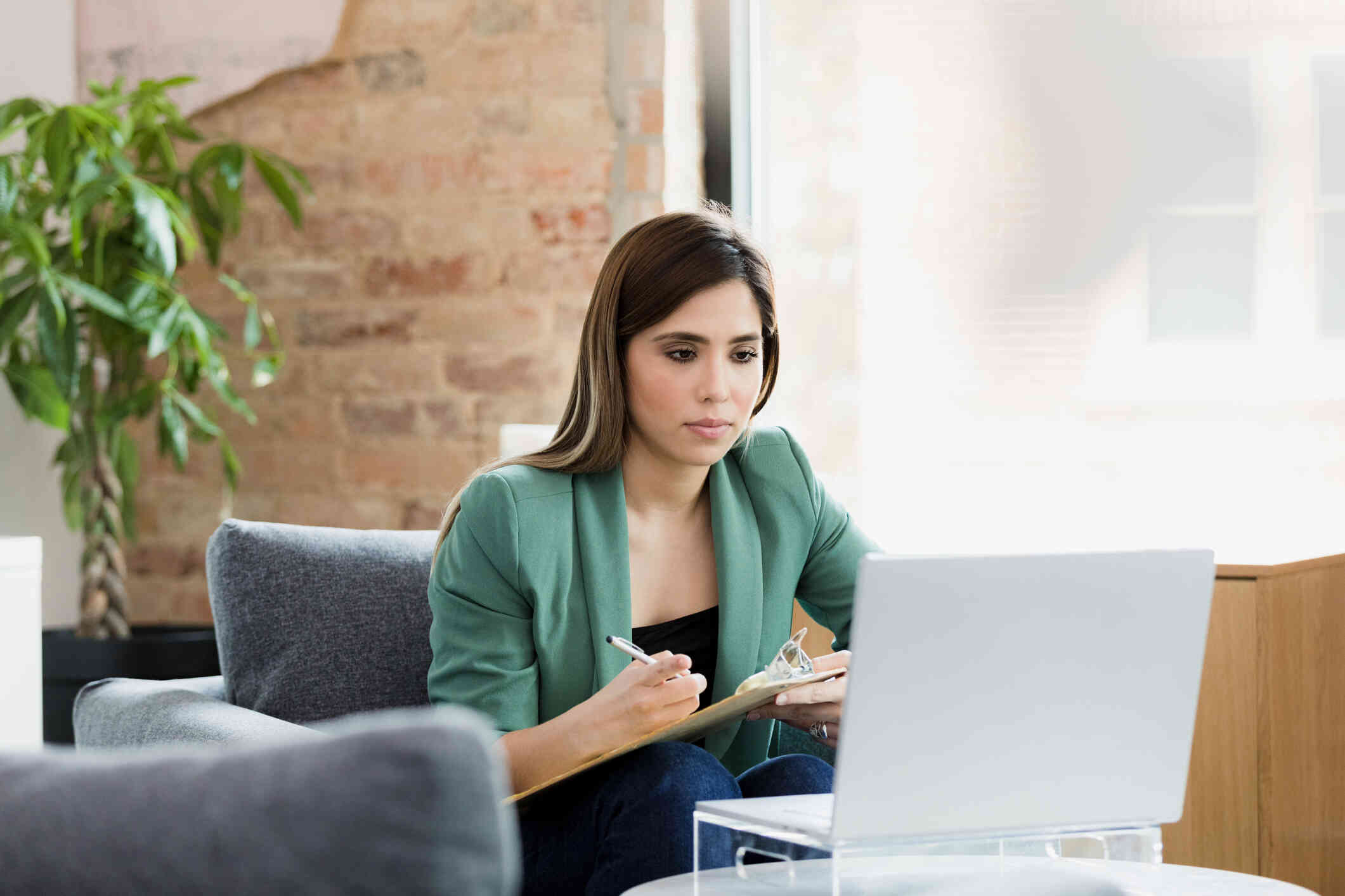 A woman in a green jacket sits in an armchair with a clipboard and pen while looking at the laptop open on a table infront of her with a serious expression.