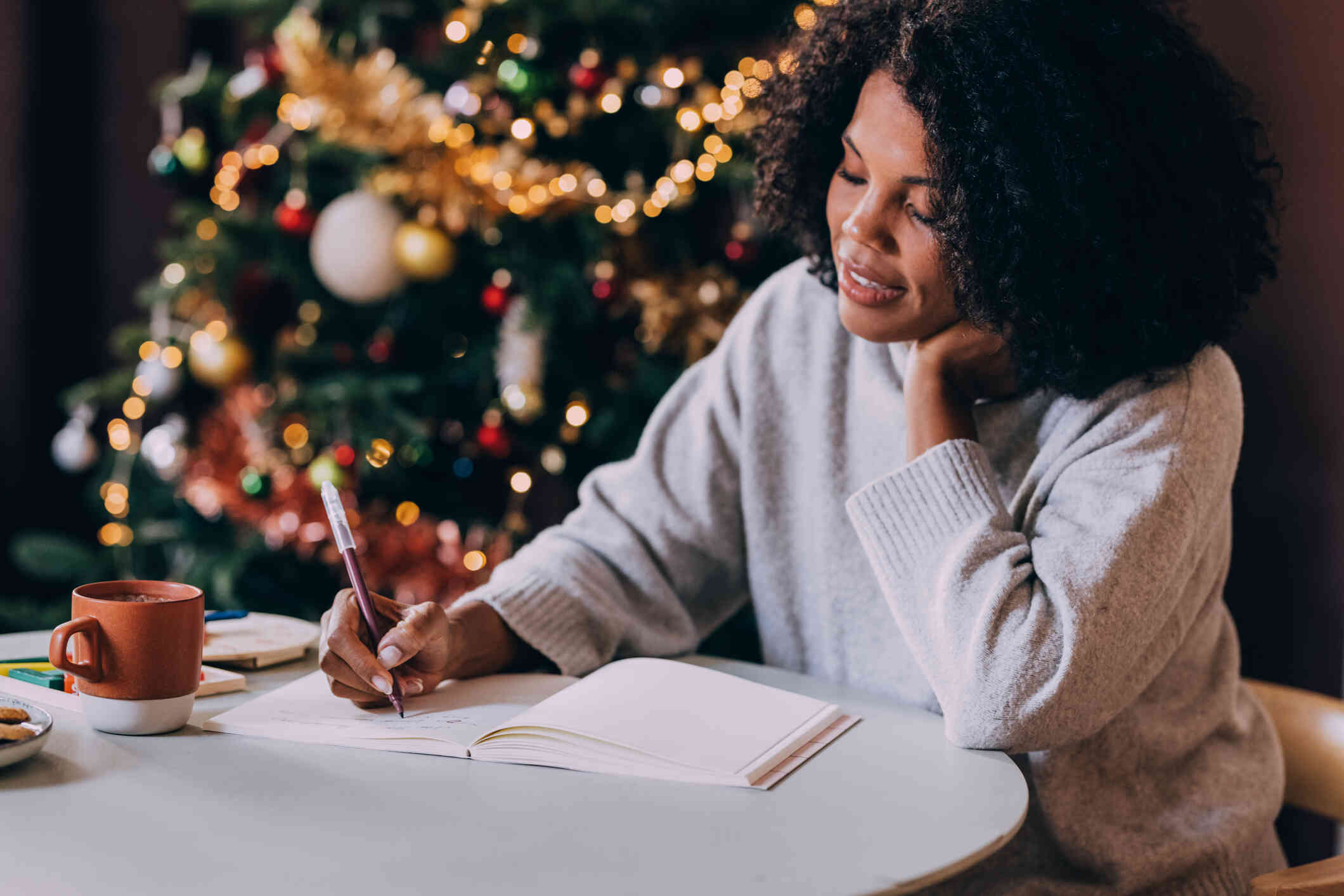 A woman sits at a table next to a christmas tree while writting in a notebook.