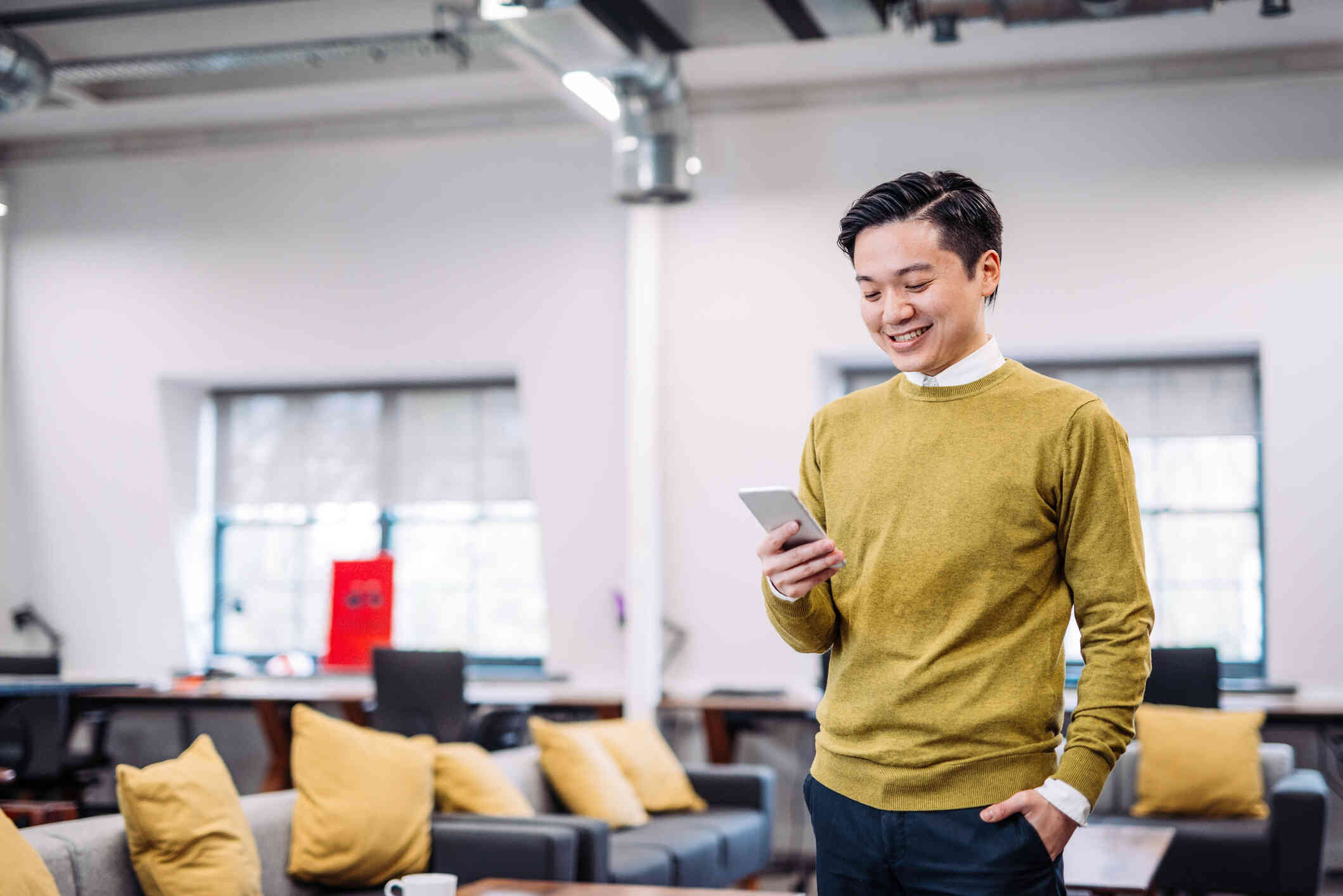 A man in asweater stands in a large room while looking down at the phone in his hand with a smile.