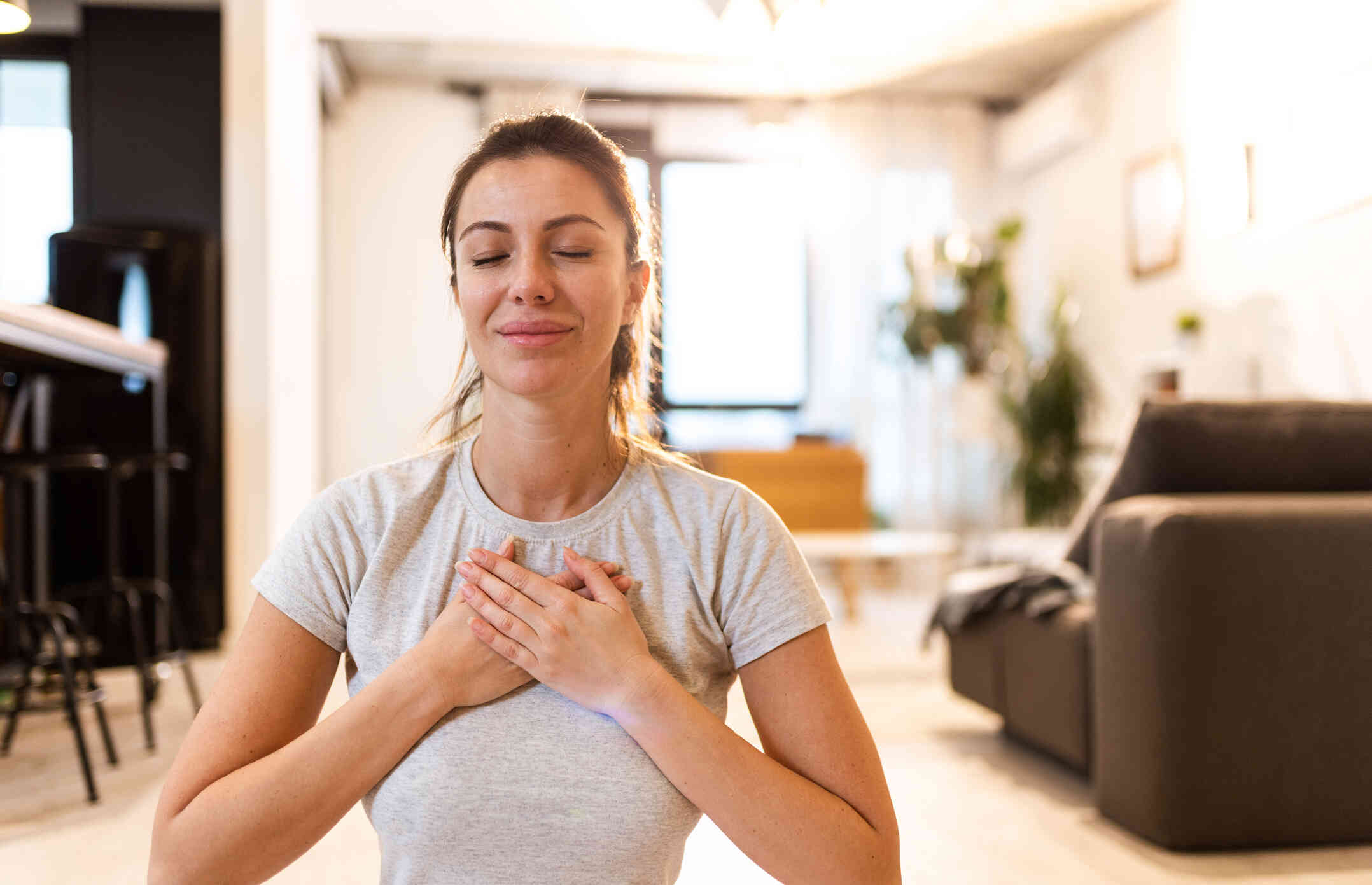 A woman in a white shirt sits in her home and places her hands over her heart with her eyes closed while practicing mindfulness.