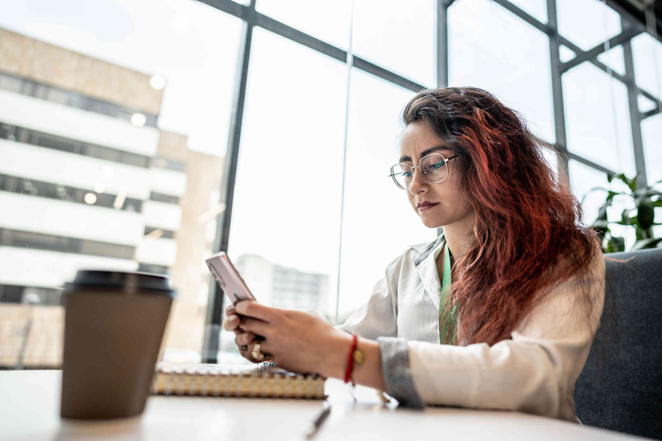 A woman with glasses sits at a table near a large window and looks at the cellphone in her hand.