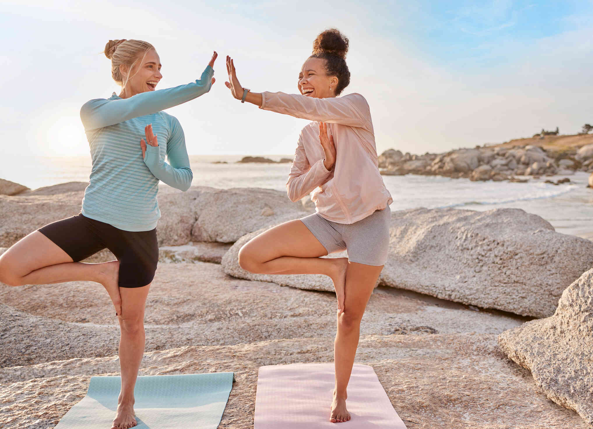 Two woman smile while high-fiving as they do yoga together on a beach.