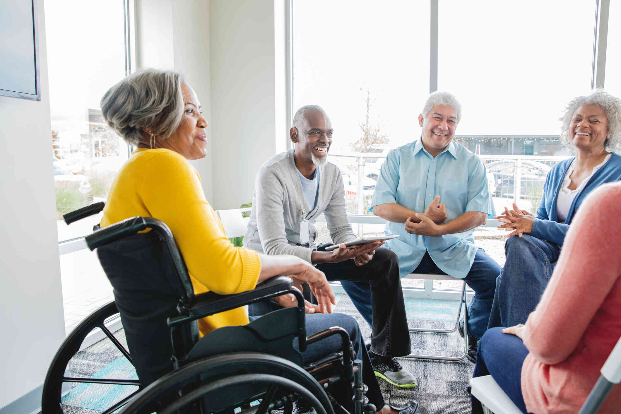 A group of elderly adults sit in a group therapy circle with a male therapist as they all talk and smile.