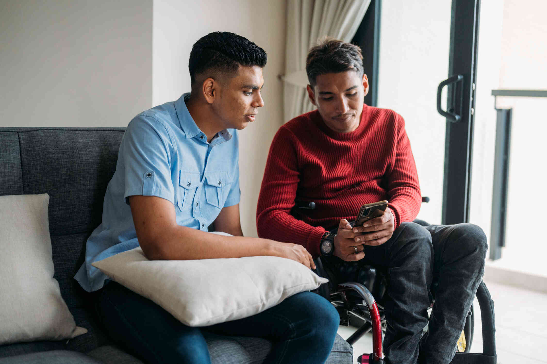 A man in a wheelchair leans over to show a male relative sitting next to him some information on the phonein his hand.