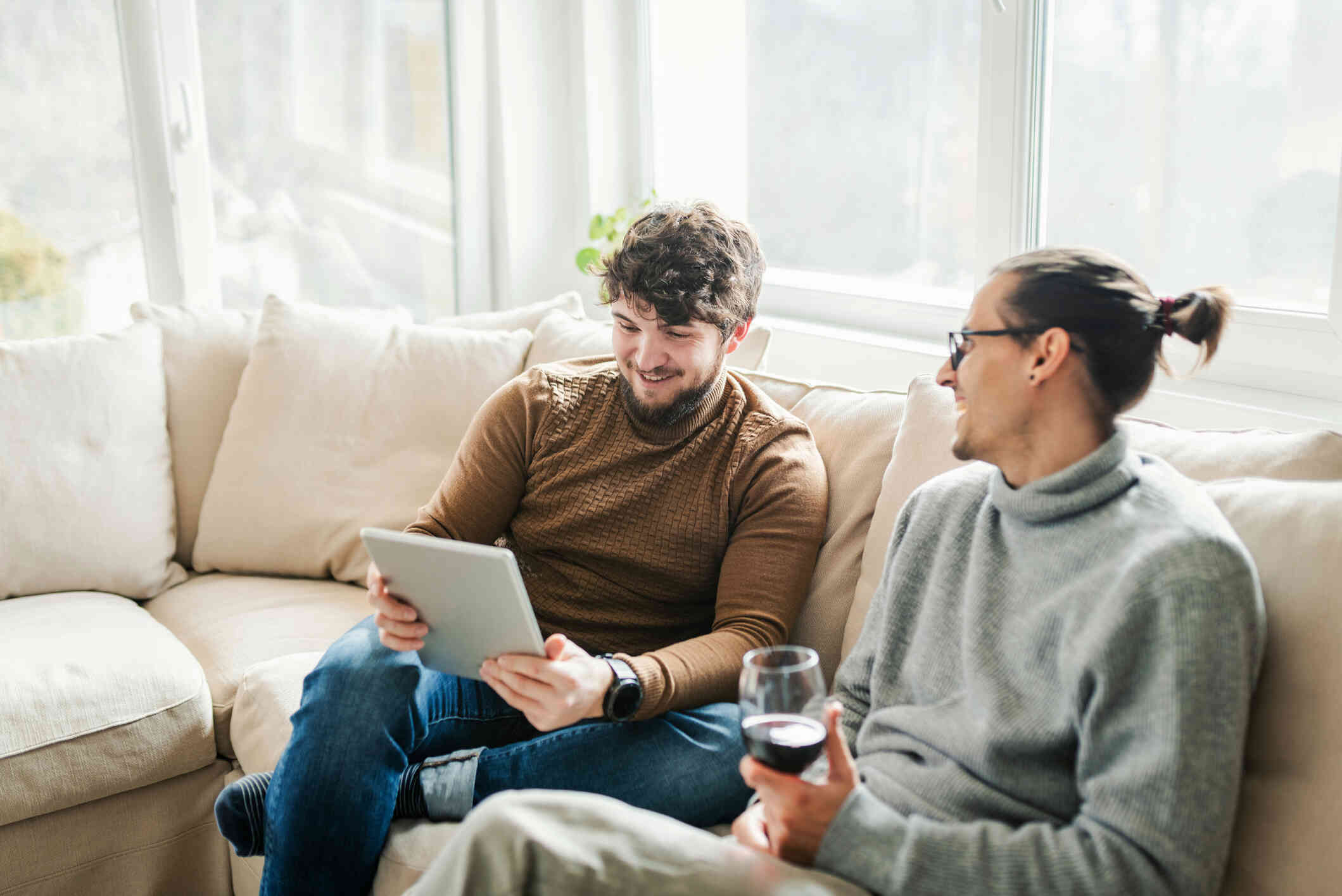 A male couple sit next to each other on the couch as one man looks at the tablet in his hand and the other holds a glass of wine.