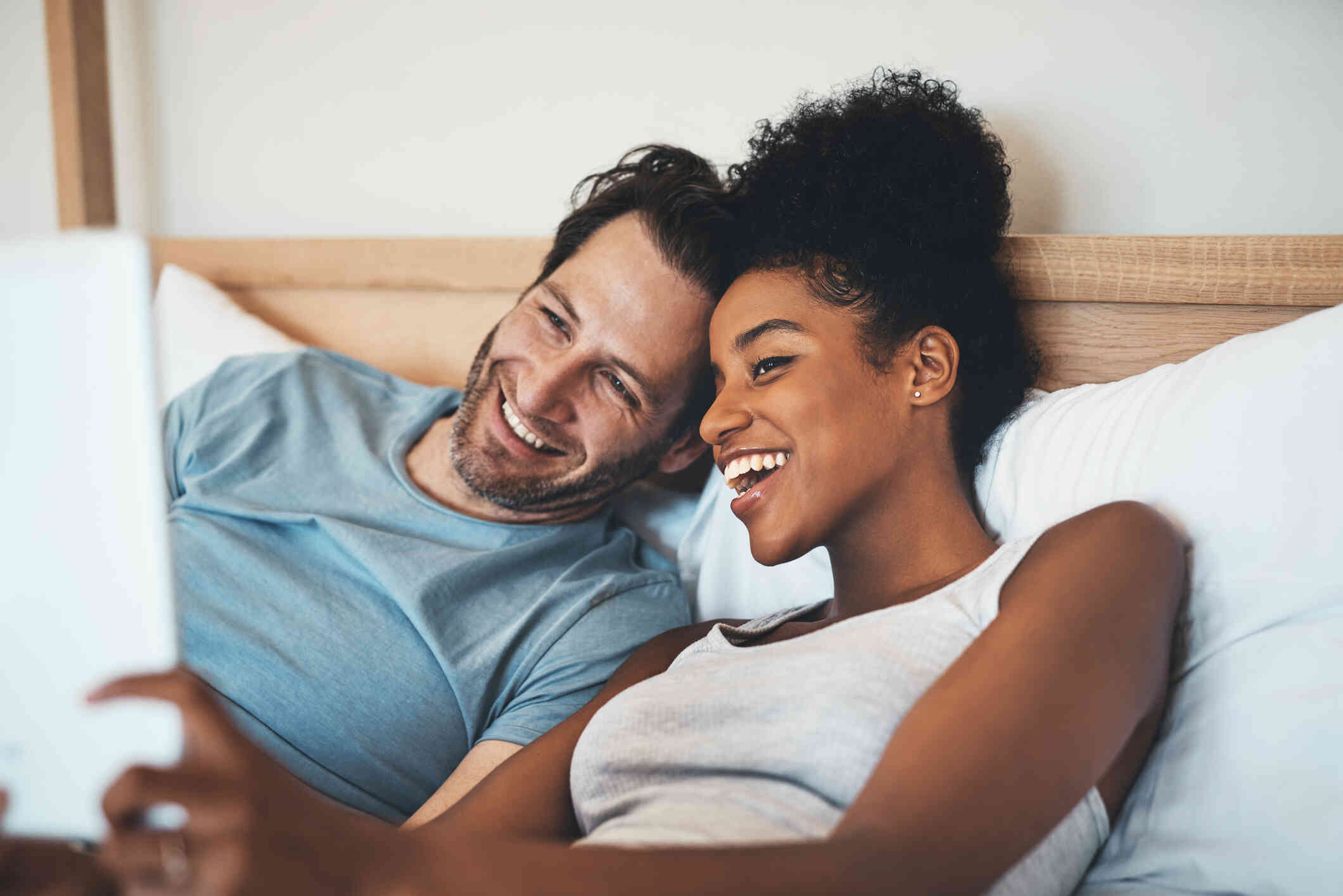 A male and female couple sit up in bed with their backs agains the headboard while smiling and taking a selfie together.