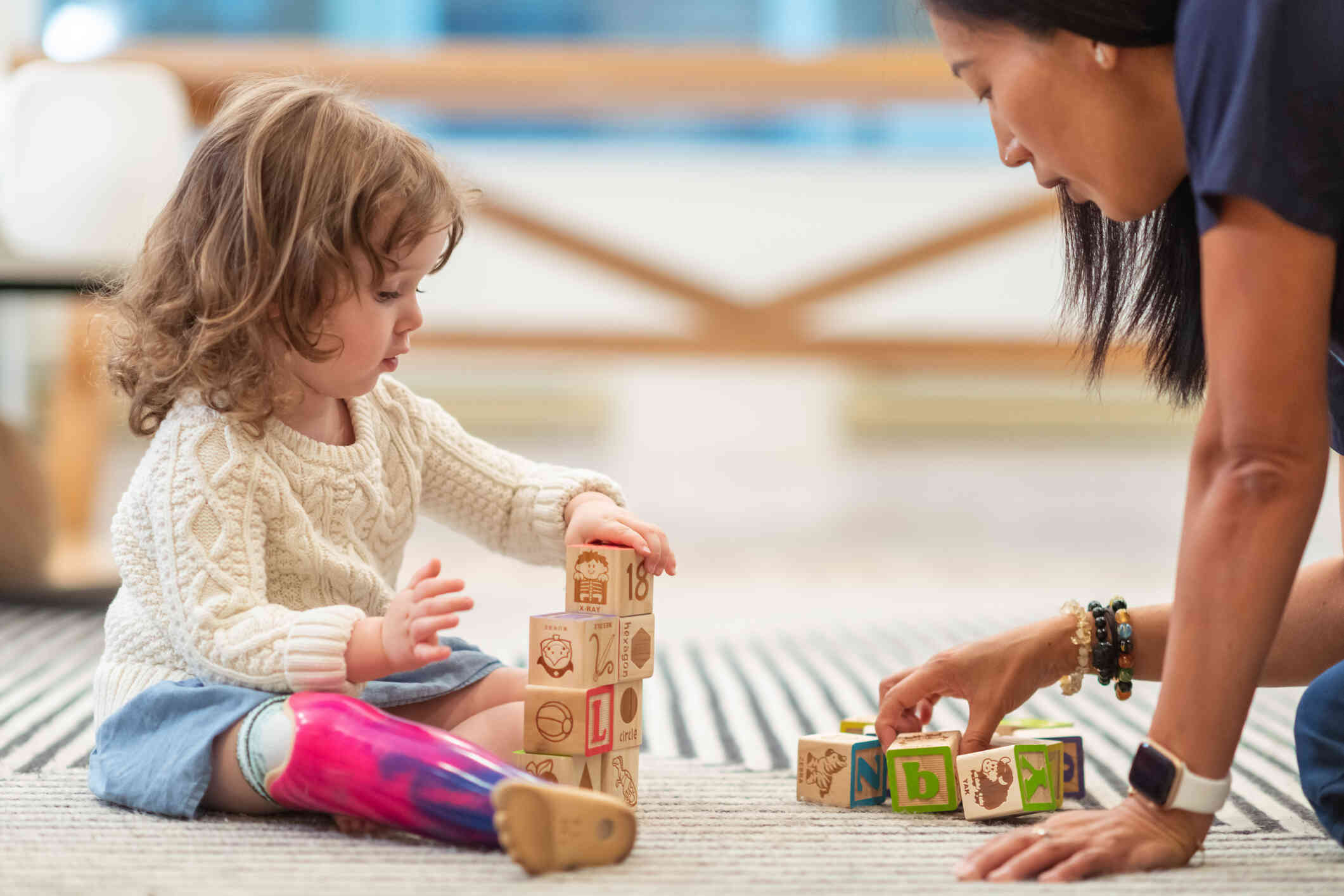 A young girl with a prosthetic leg sits on the floor playing with blocks as a woman with long dark hair sits across from her and speaks to her.