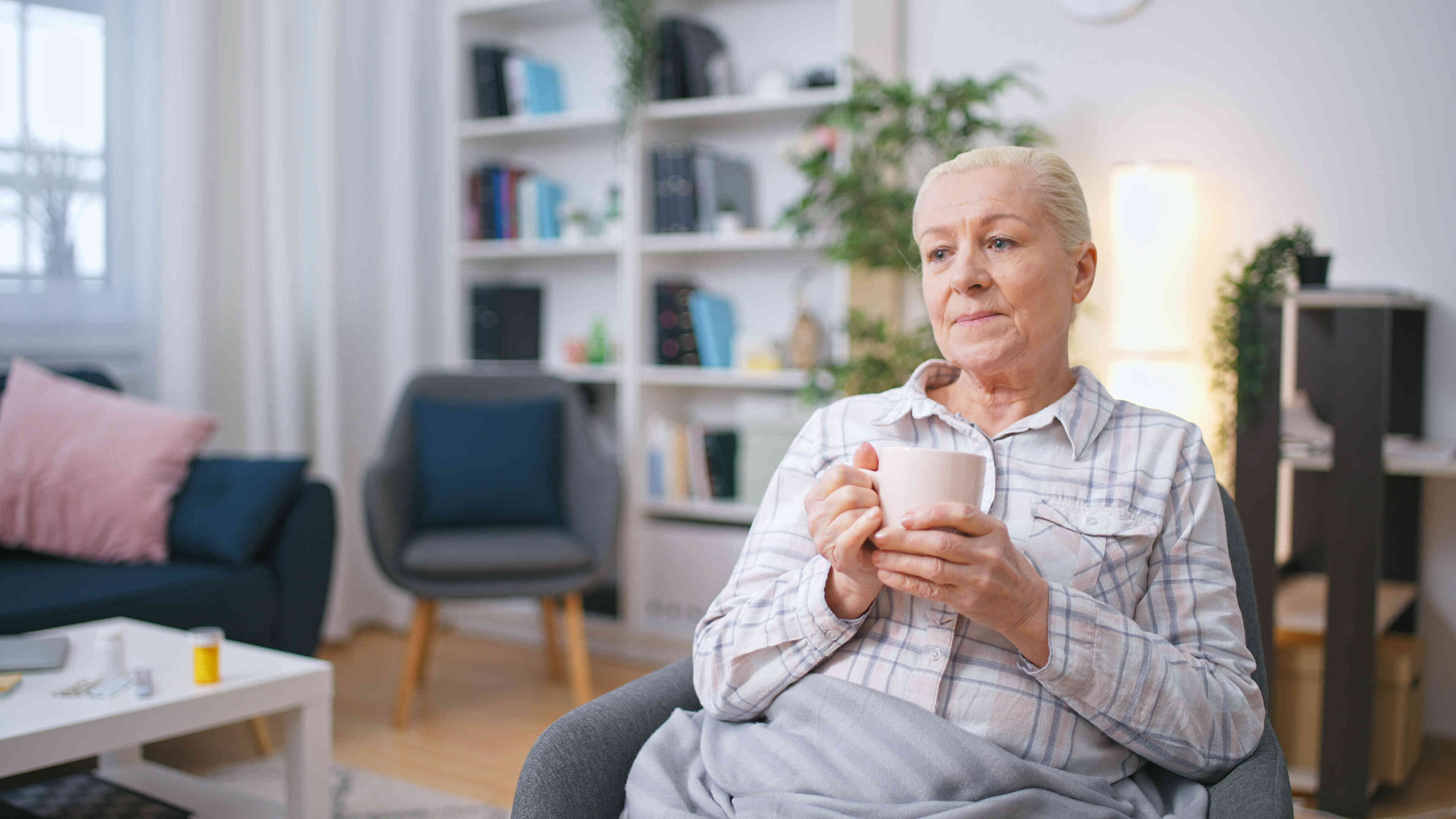 A mature woman in a plaid shirt sits in an armchair in her home and holds a coffee mug while gazing off with a worried expression.