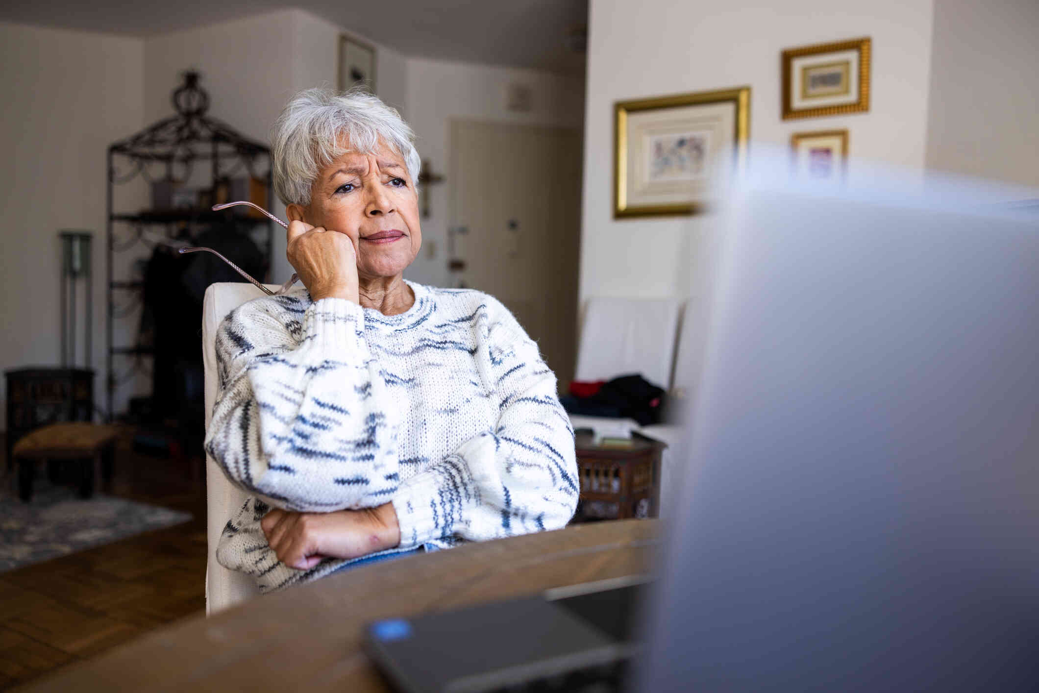 An elderly woman sits at a table with her tablet infront of her and holds her glasses while looking at the tablet with a worried expression.