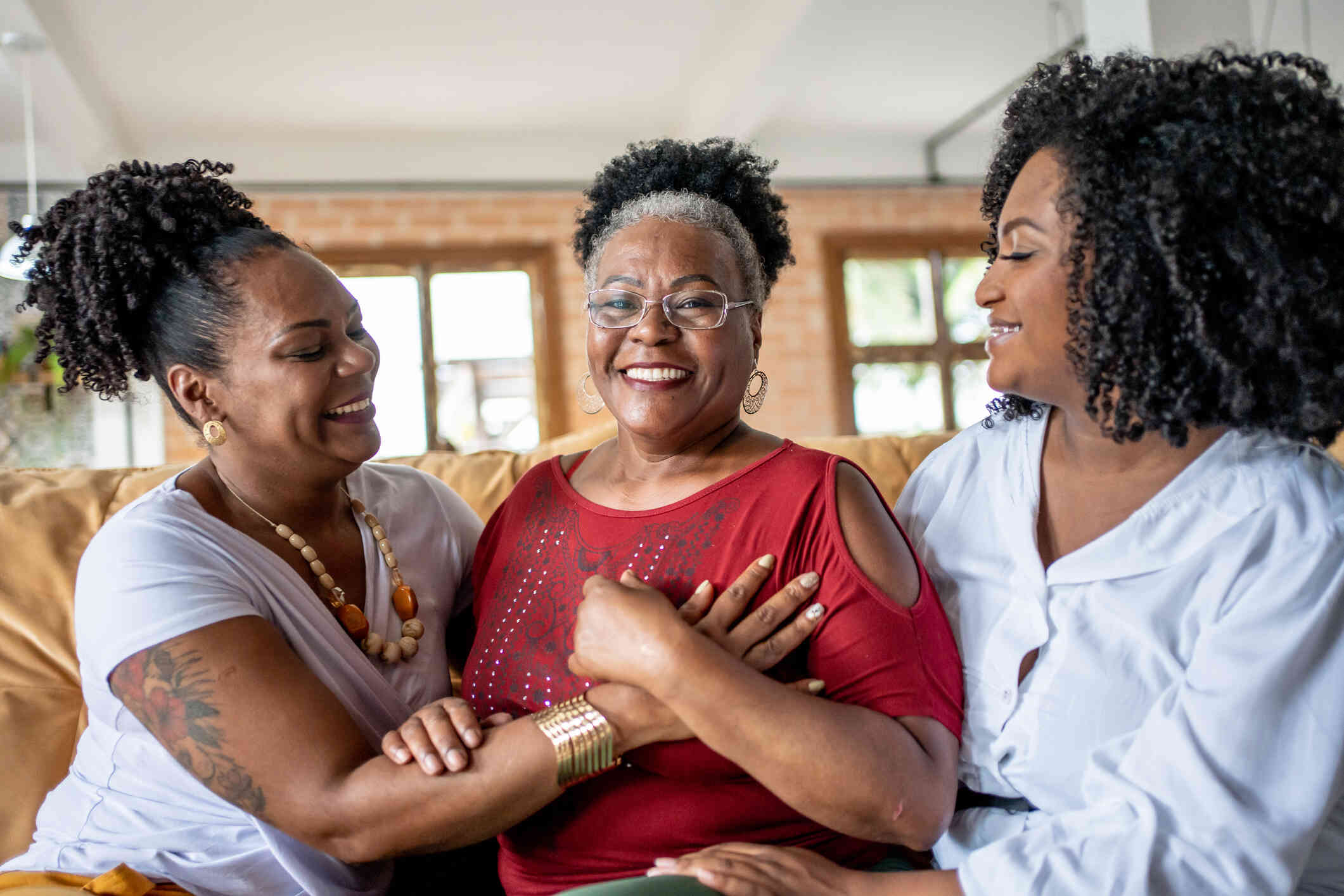 Three adult female family members embrace one another while smiling and standing in a home.