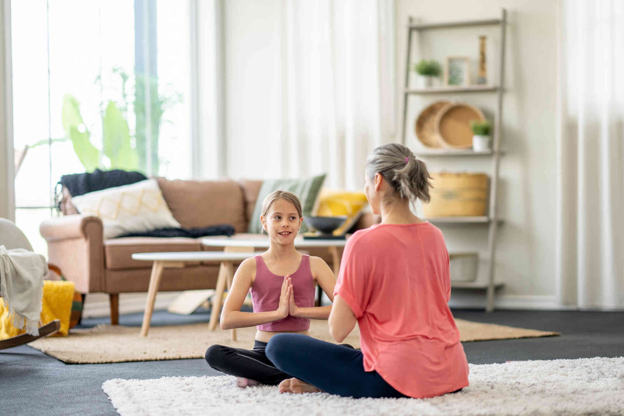 A young girl sits on the livingroom floor across from her mother as they practice yoga together.