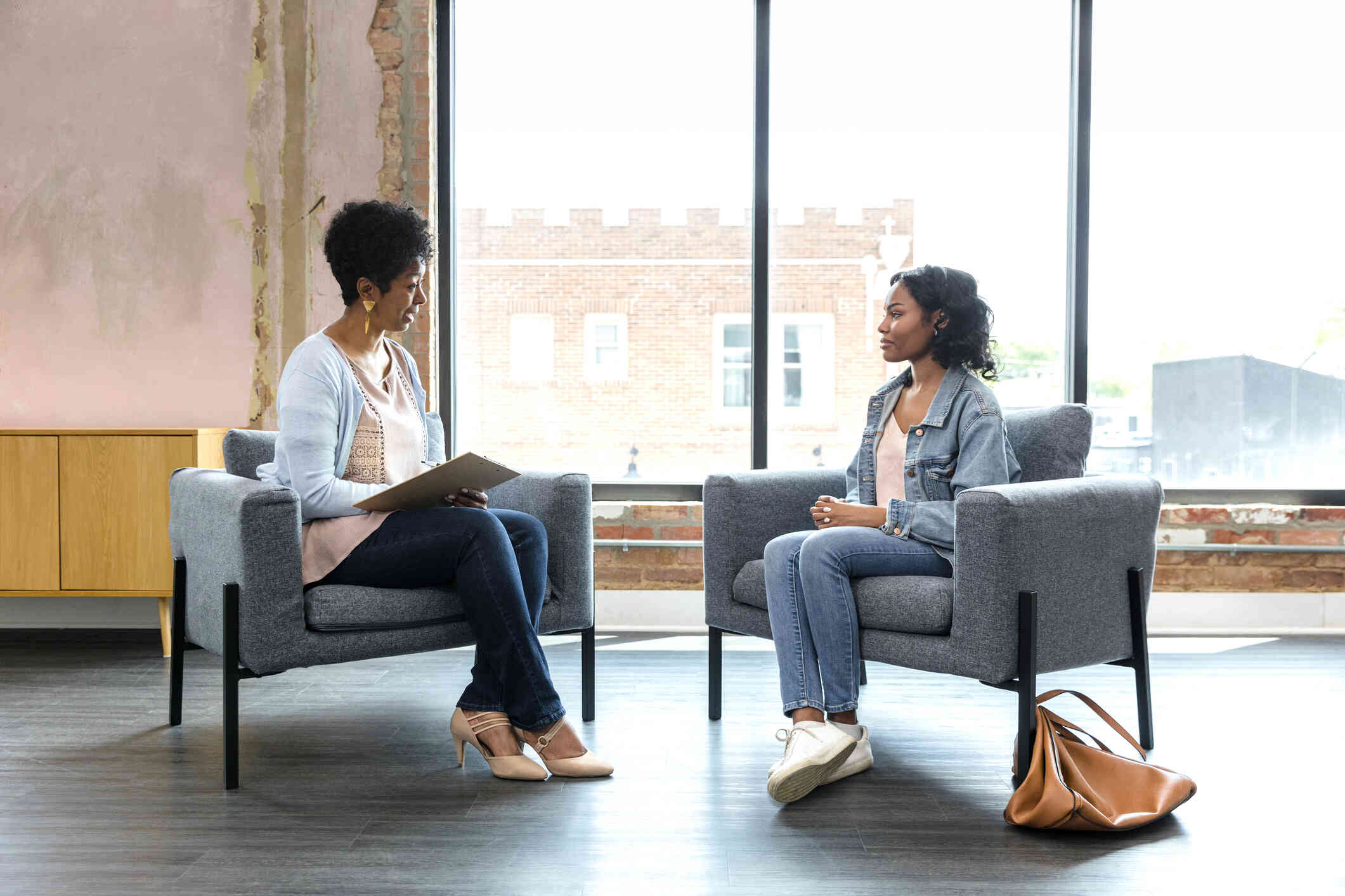 A woman in a blue jean jacket sits across from her female therapist as she talks during a therapy session.