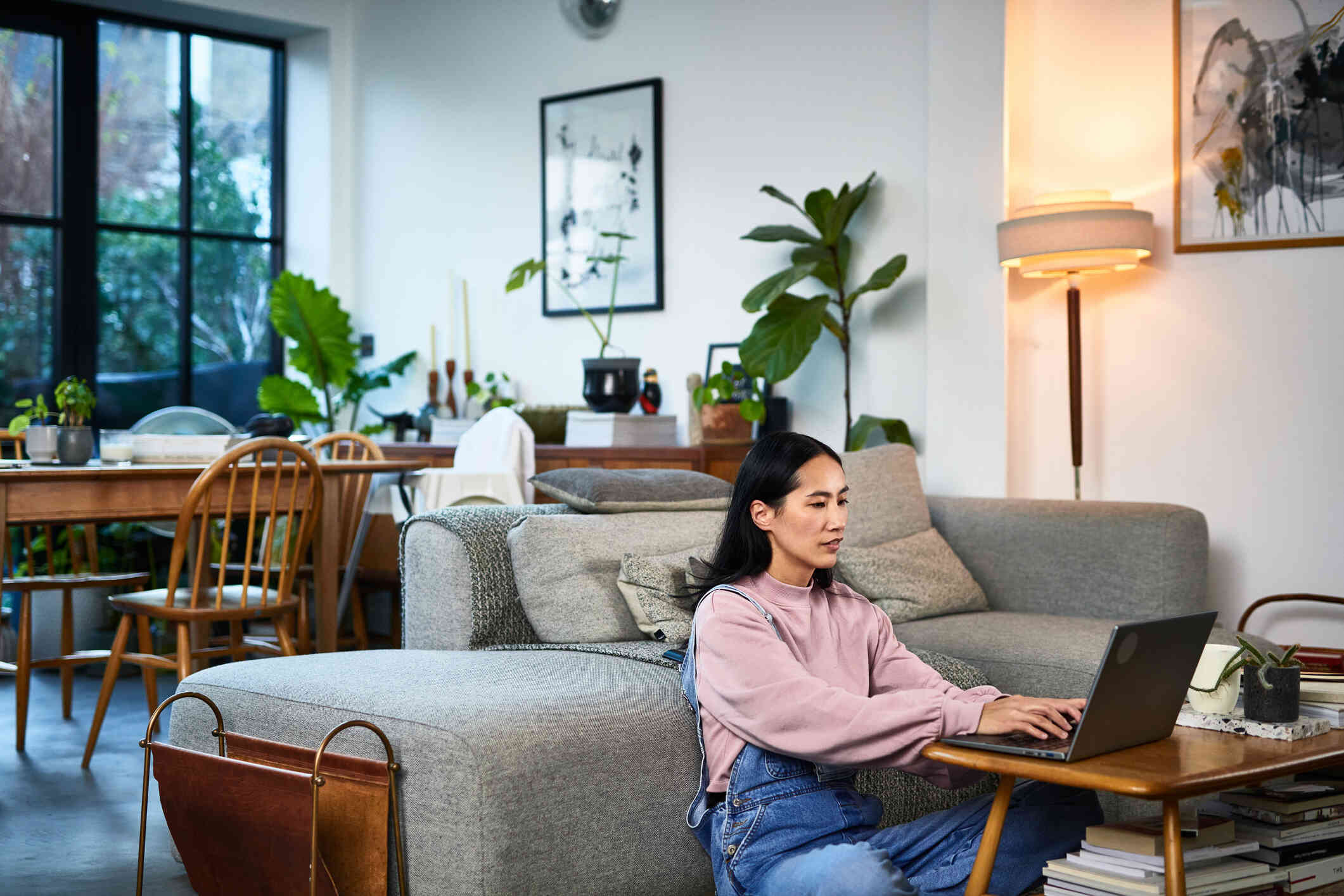 A woman in a pink sweater sits on the living room floor with her back against the couch as she types on the laptop on the coffee table infront of her.