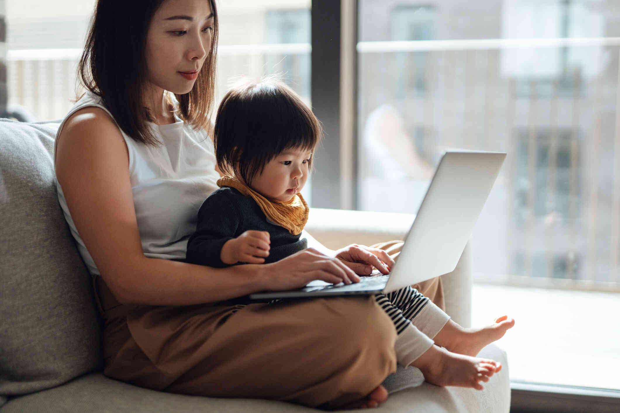 A woman sits in a cair in her home mear a window with her toddler and laptop in her lap as she types on the keyboard.