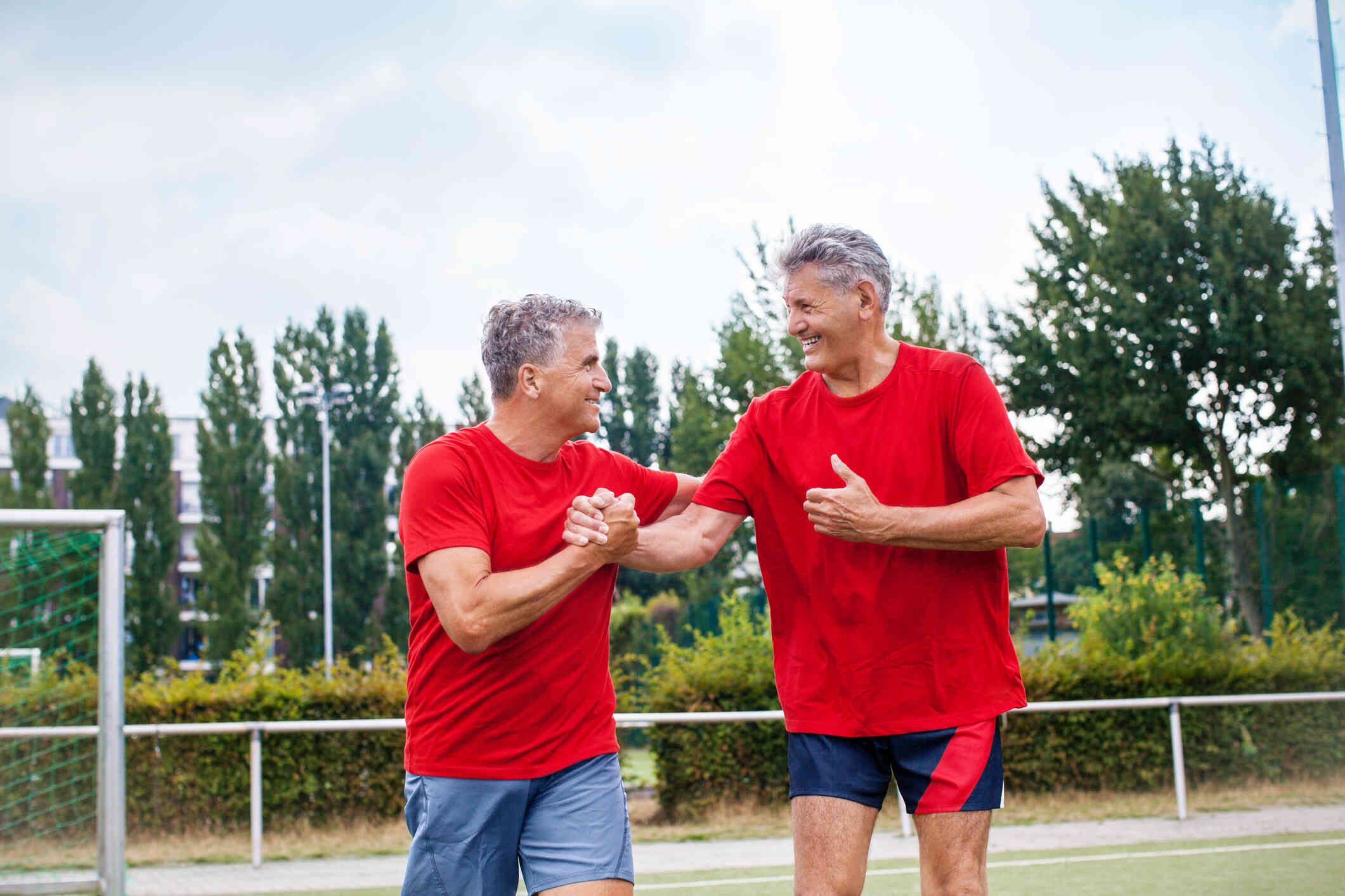 Two mature men wearing red t-shirts smile as they clasp hands and pat each other on the back while exercising outside.