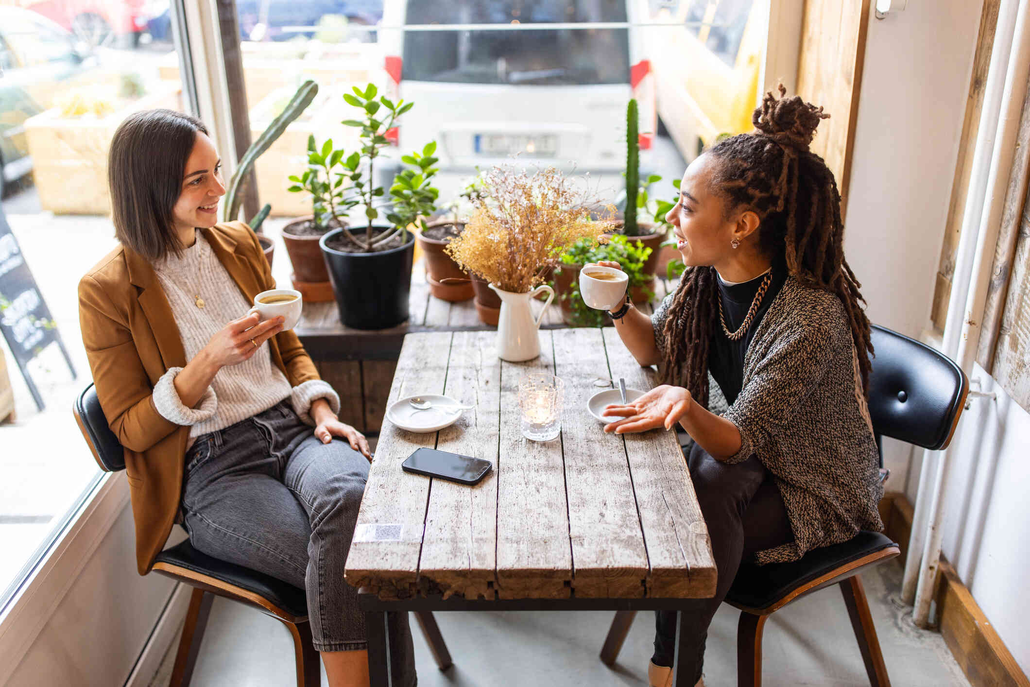 Two woman sit across from each other at a table in coffee shop and chat while drinking their coffee.