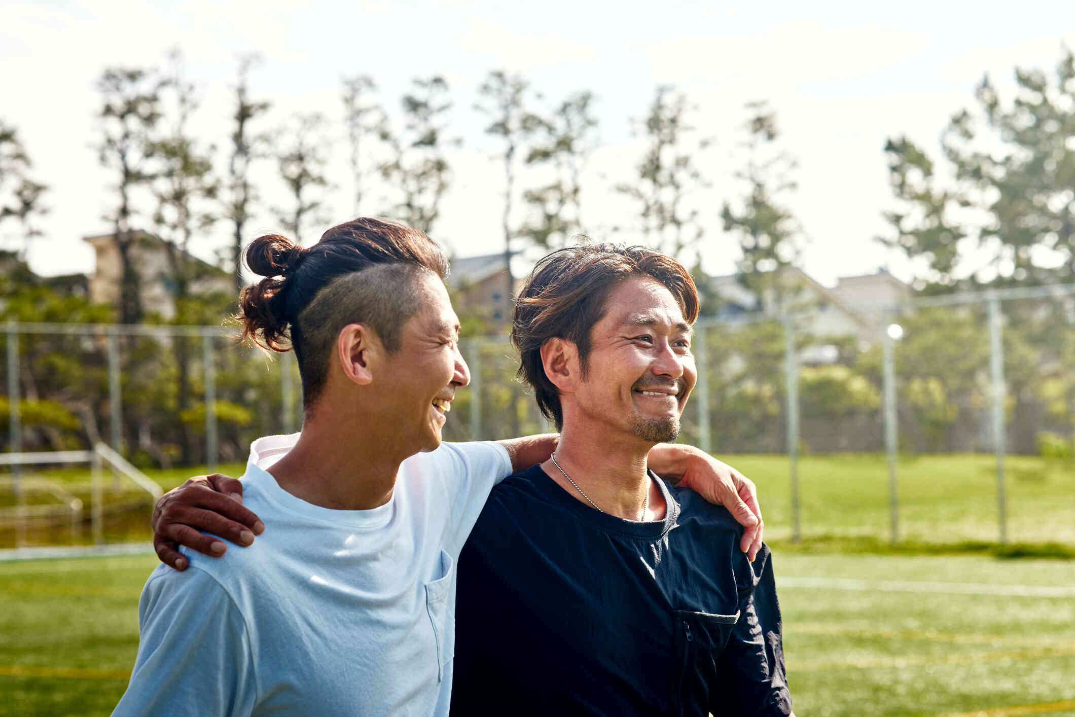 A close up of two men as they wrap their arms around each others shoulders and smile while walking outside on a sunny day.