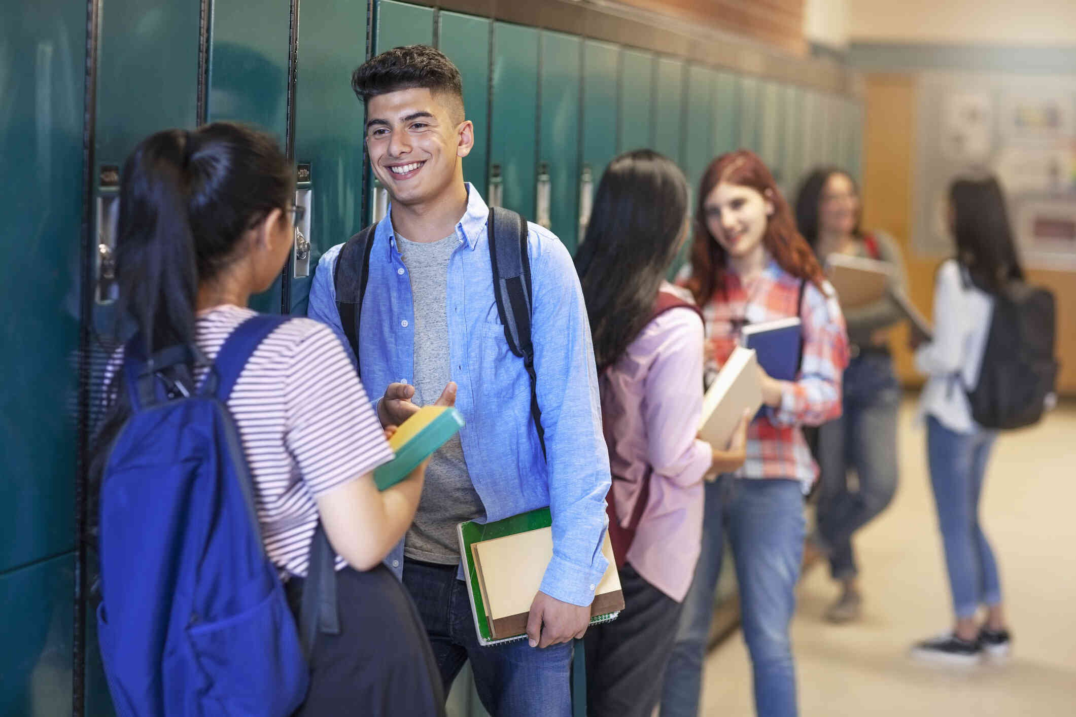 A teenage boy wearing a backpack smiles at a female friend as he leans against a row of blue lockers in a crowded school hallway.