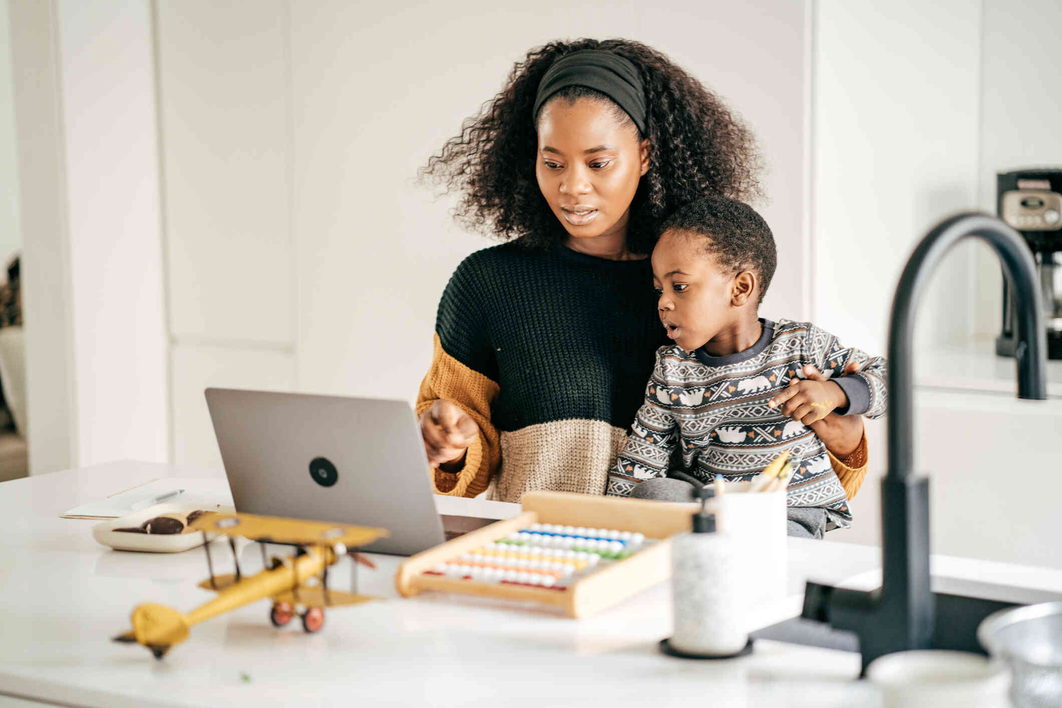 A woman wearing a headband and a sweater holds a young boy on her lap and points to something on the screen of a laptop which is sitting on the counter in front of her surrounded by toys.