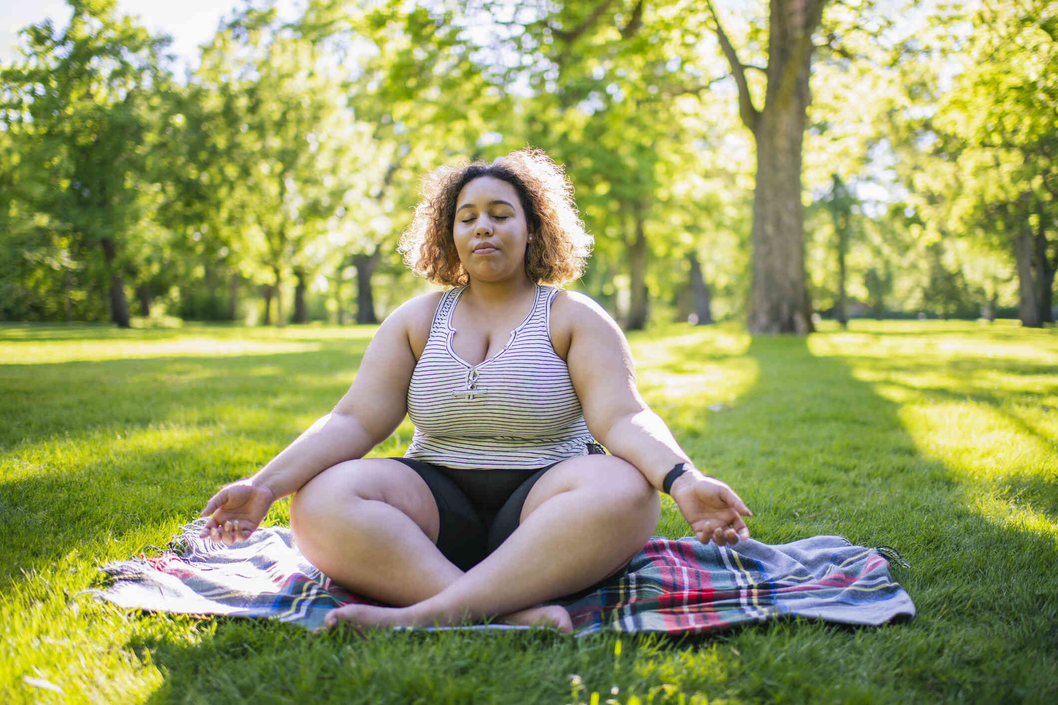 A woman sitting cross-legged on blanket in a park, meditating with her eyes closed.