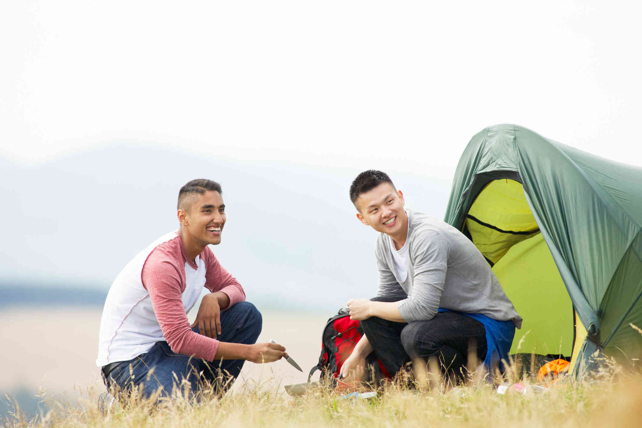 Two male friends set up a camp site together in an open field while smiling.