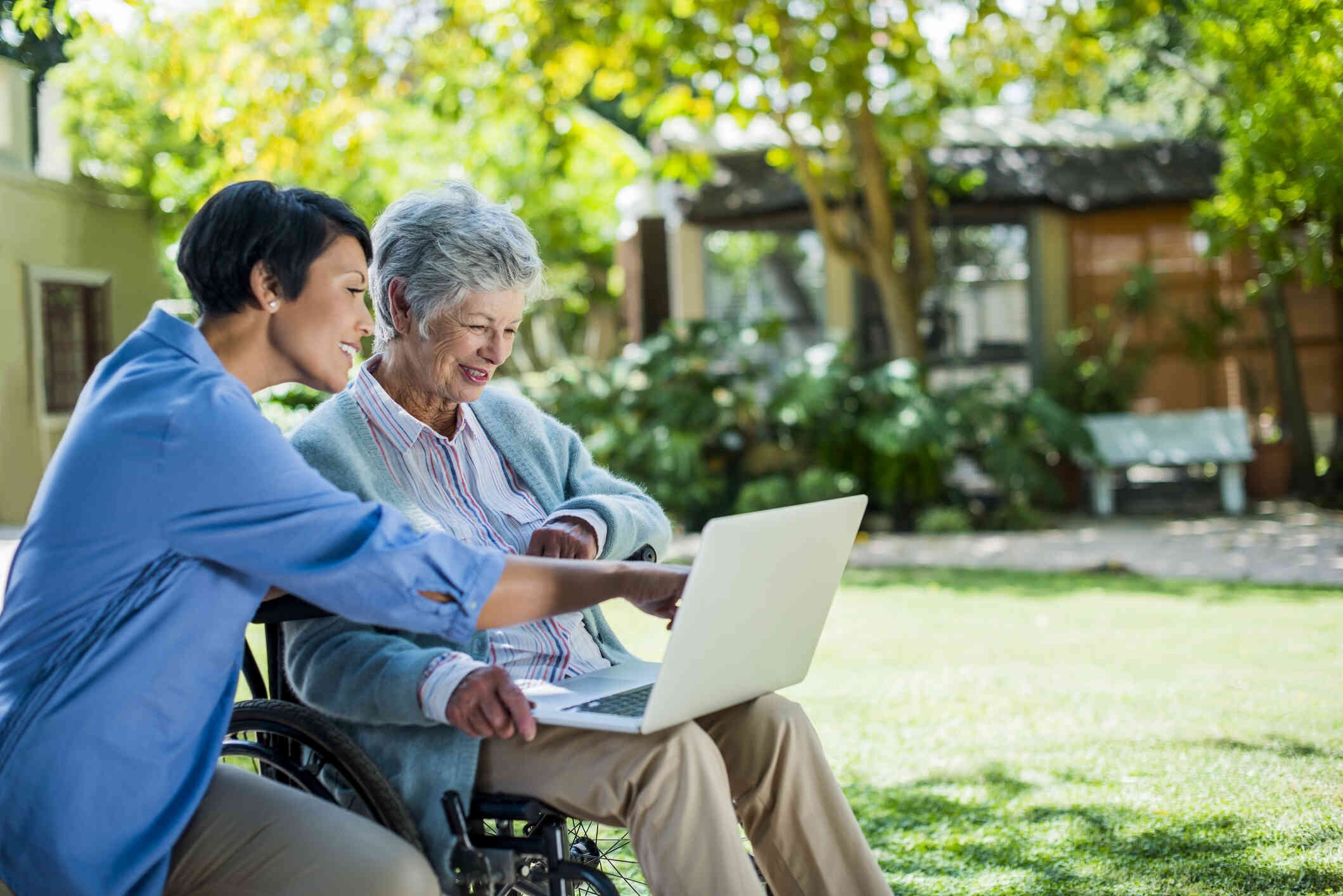 An elderly woman in a wheelchair sits outside with a laptop open in his lap as a woman next to her points to some information on the screen.