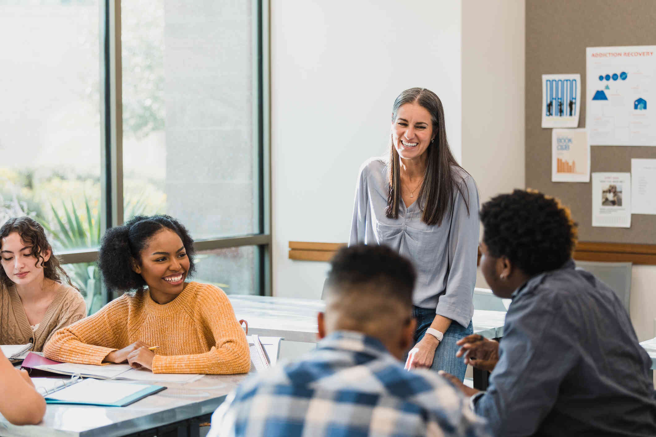 A woman stands in a classroom and smiles as she looks at a group of male and female students who are sitting at tables with binders open on them.