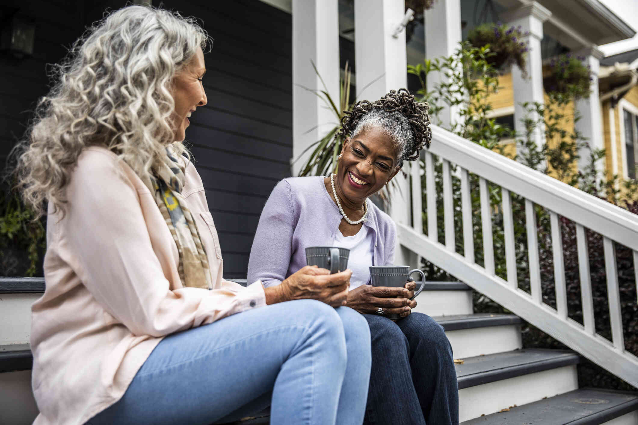 Two mature female friends sit next to each other on the front porch steps while laughing and holding cups of coffee.