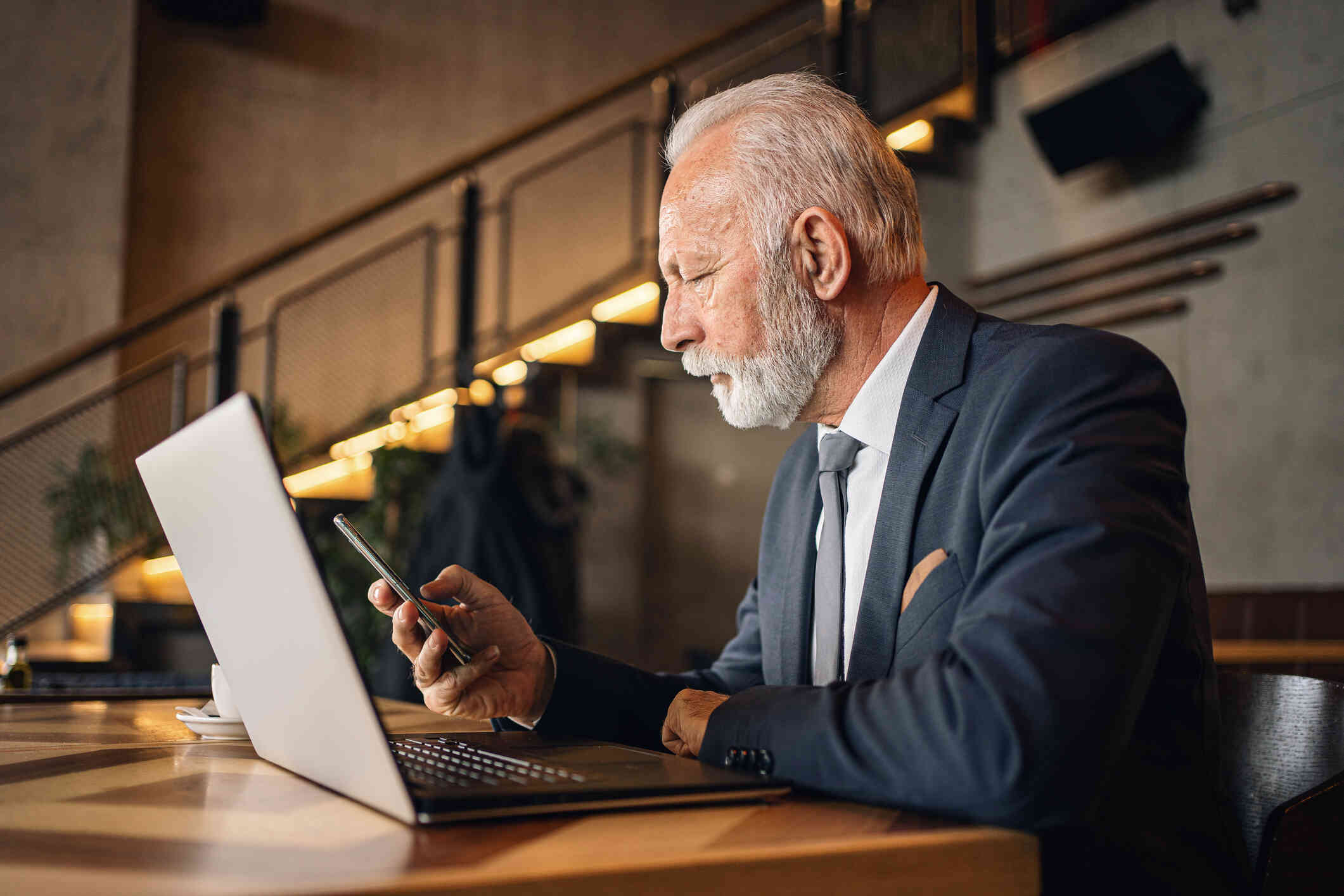 An elderly man in a suit sits at a table with his laptop open infront of him as he looks at the phone in his hand.