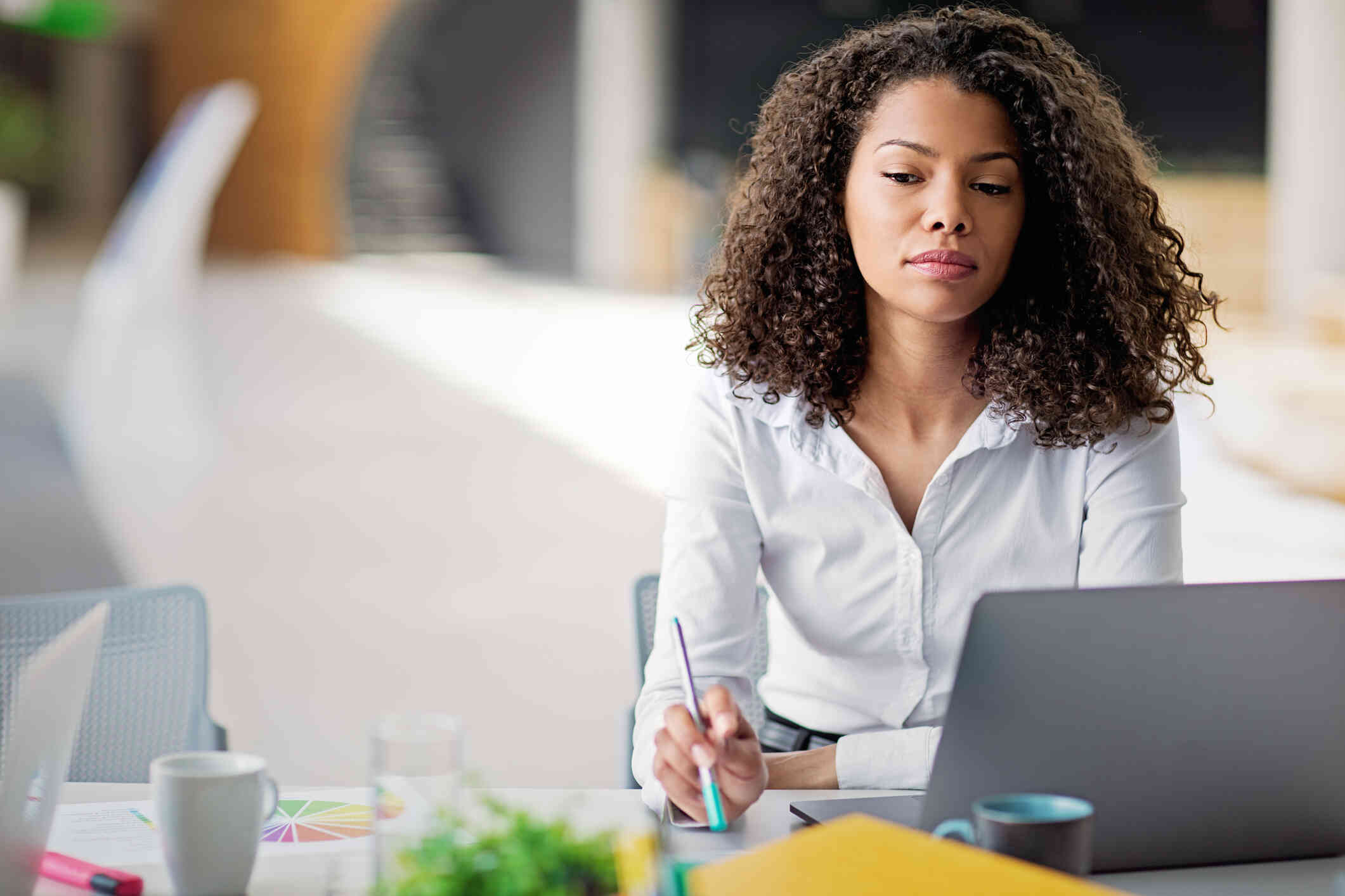A woman in a white buttin down shirt sits at a table with her laptop and gazes off while deep in thought.