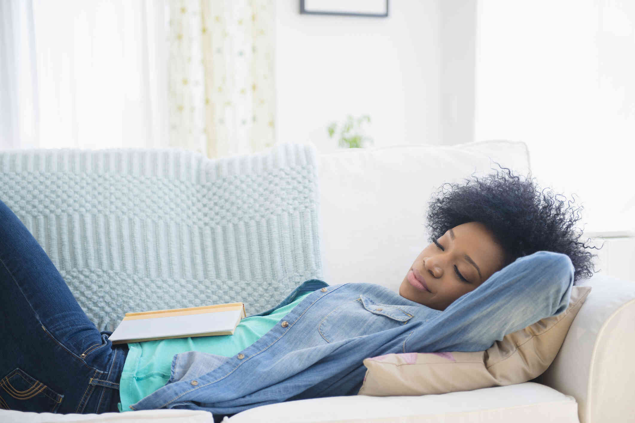 A woman ina blue jean jacket lays asleep on her couch during the day with an open book resting on her stomach.
