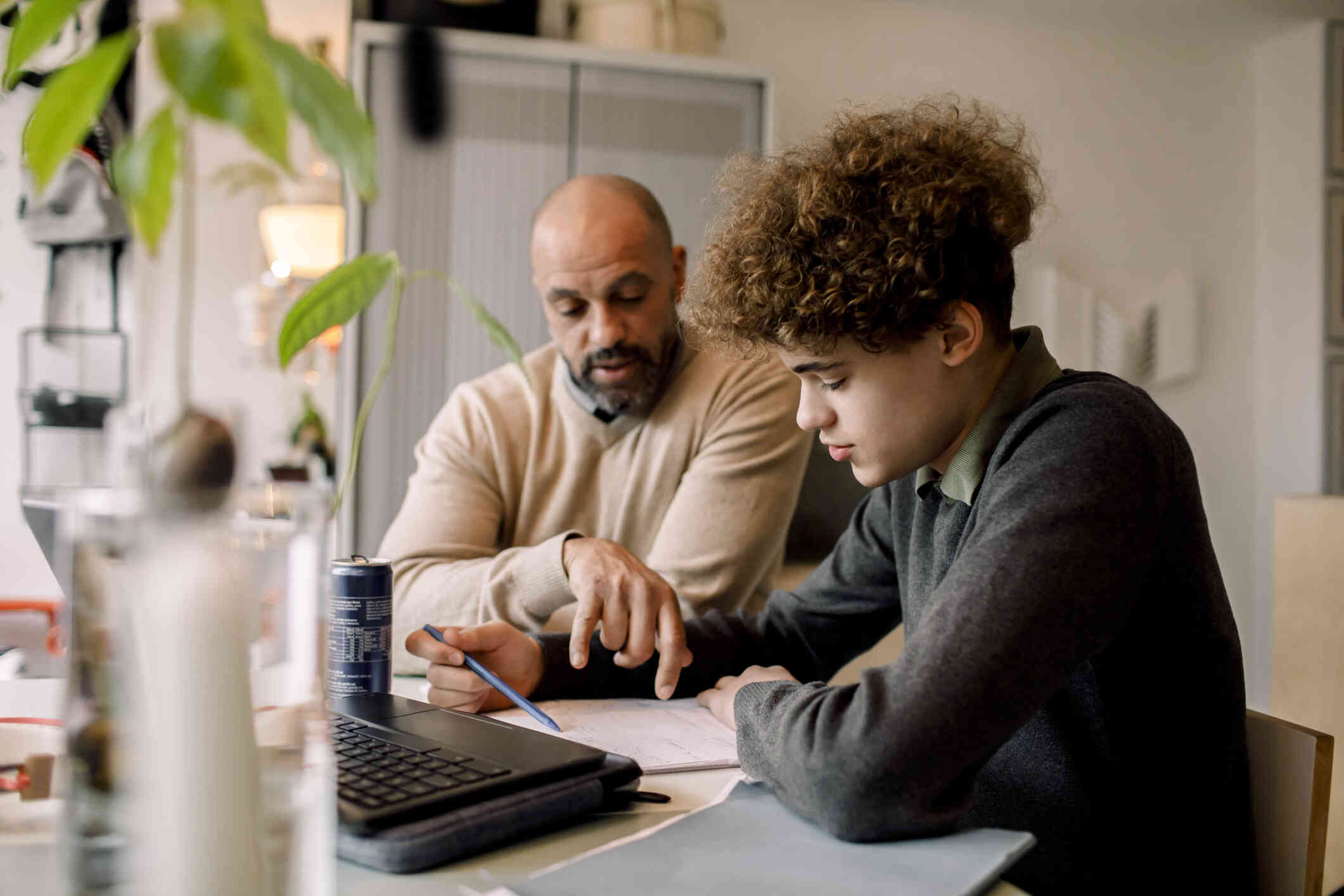 A teenage boy and his father sit together at a desk, focused on filling out documents in front of the laptop.