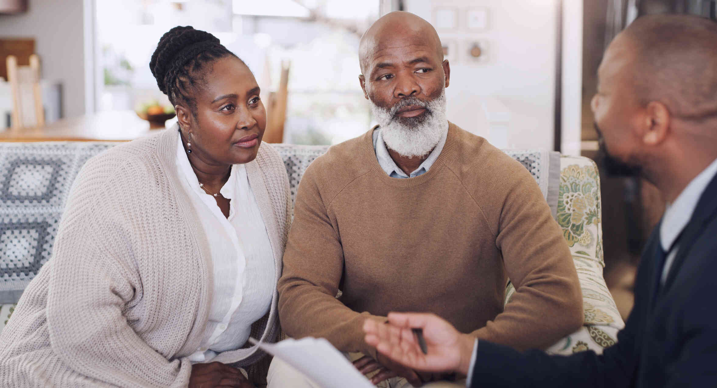 A mature man with a gray beard has a serious expression as he sits next to a mature woman wearing a tan cardigan. A man sits next to them and speaks while holding papers.