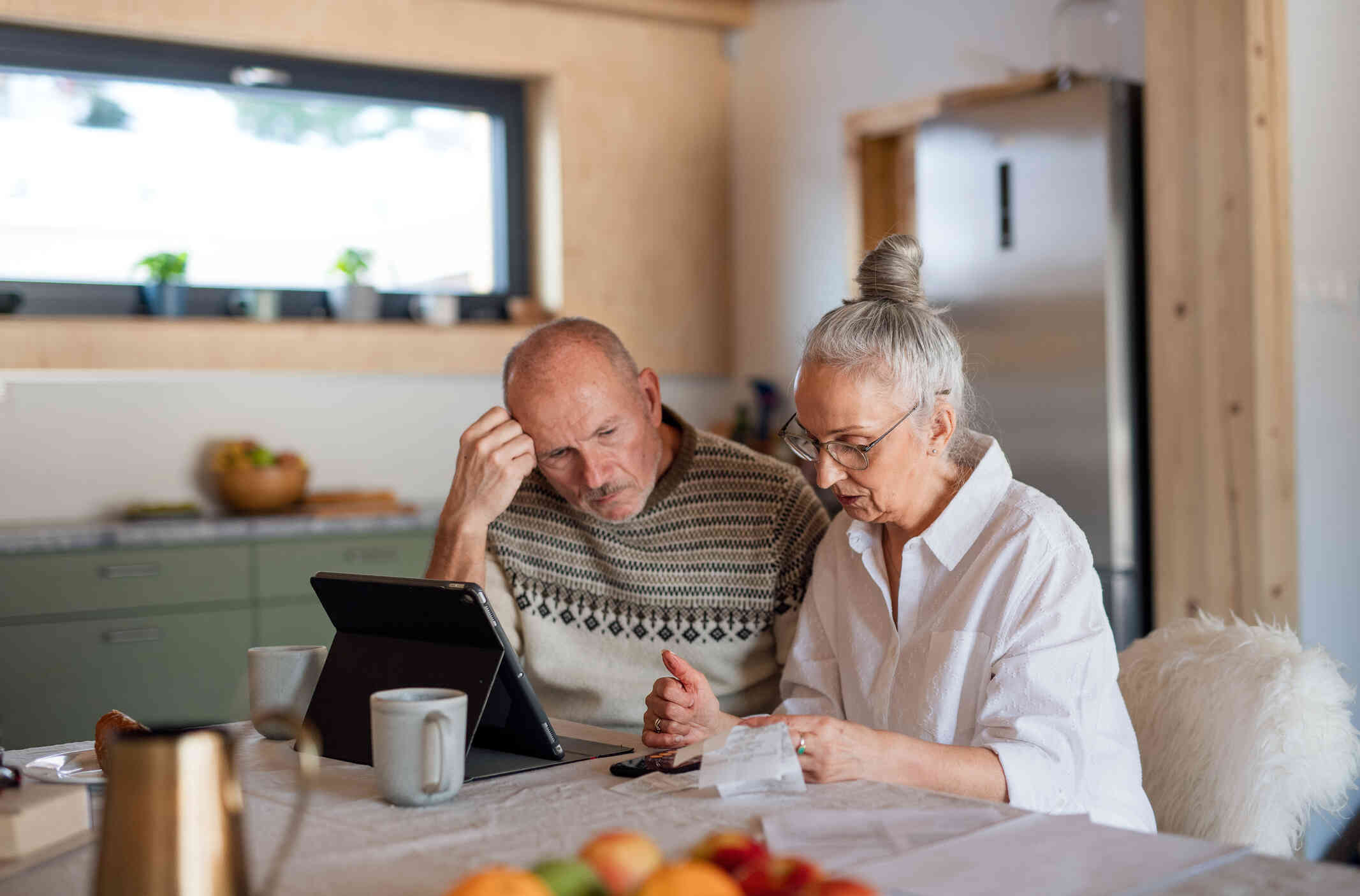 An elderly couple seated at a table appears focused, reviewing papers, with a tablet and coffee mugs nearby.